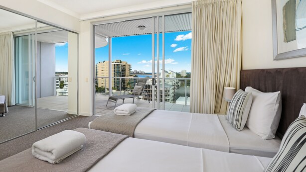 A hotel room with a large sliding glass door leading to a balcony, a bed with white linens, and a view of the cityscape. - Pandanus Mooloolaba (Mooloolaba)