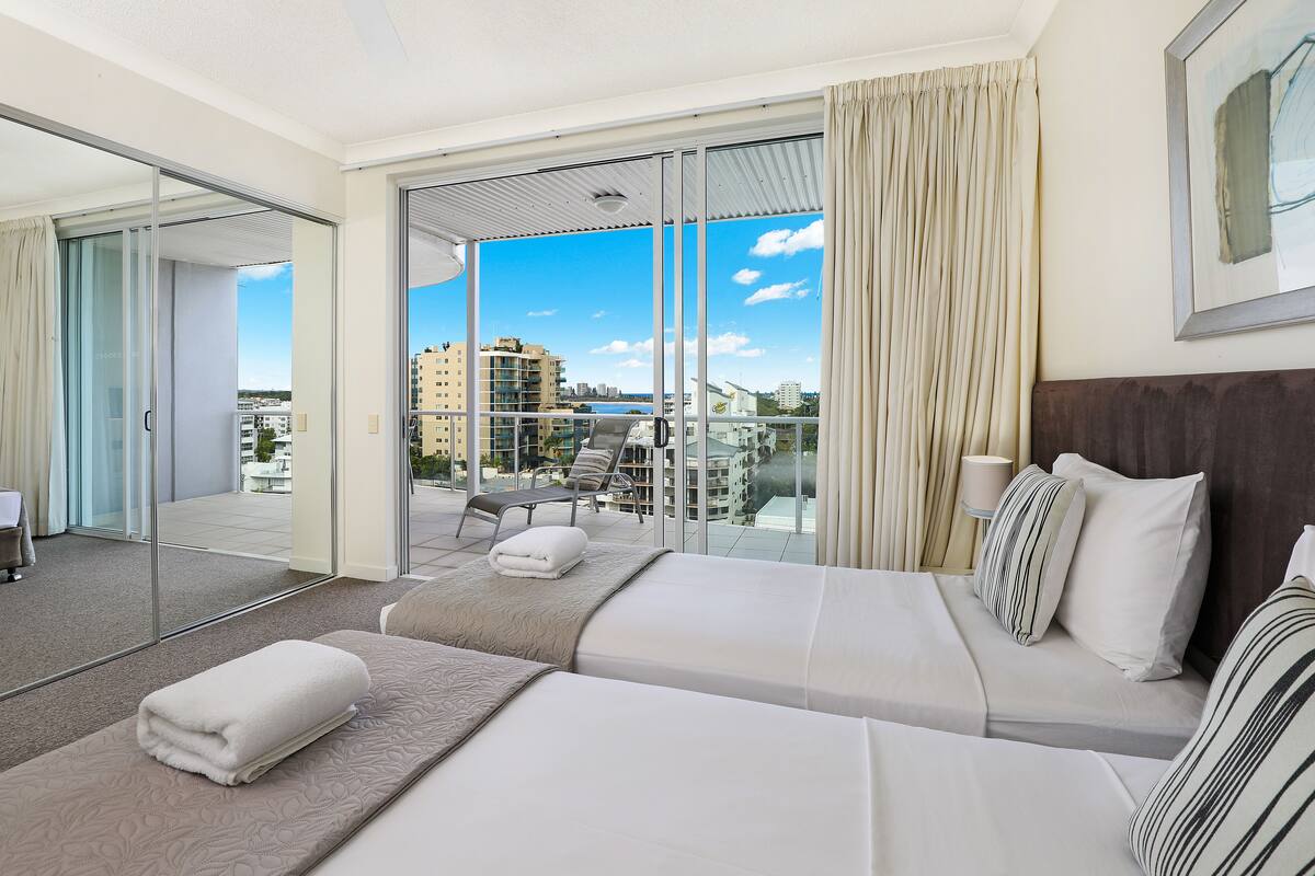Habitación de hotel con un amplio ventanal corredero que da a un balcón, una cama con ropa blanca y vistas a la ciudad.