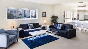 A modern living room with two black leather sofas, a white coffee table, and a blue rug. There are two side tables with lamps, a large window with a view of buildings, and a kitchen area in the background. - Pandanus Mooloolaba (Mooloolaba)
