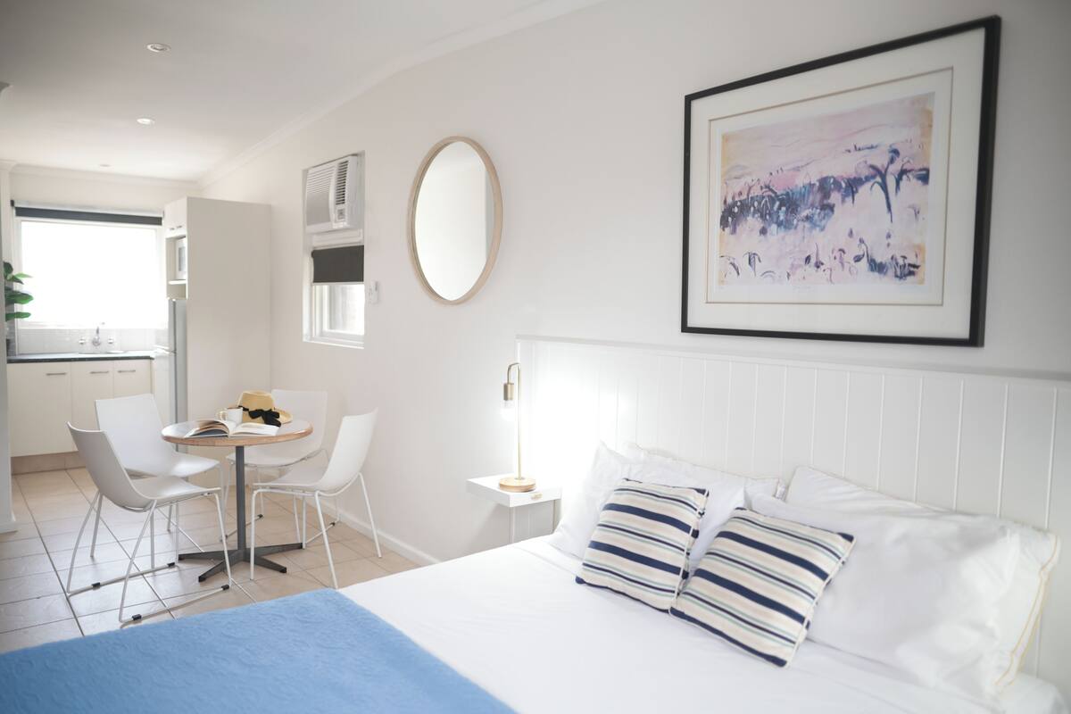 A hotel room with a bed, a desk with chairs, a window, and a painting on the wall.