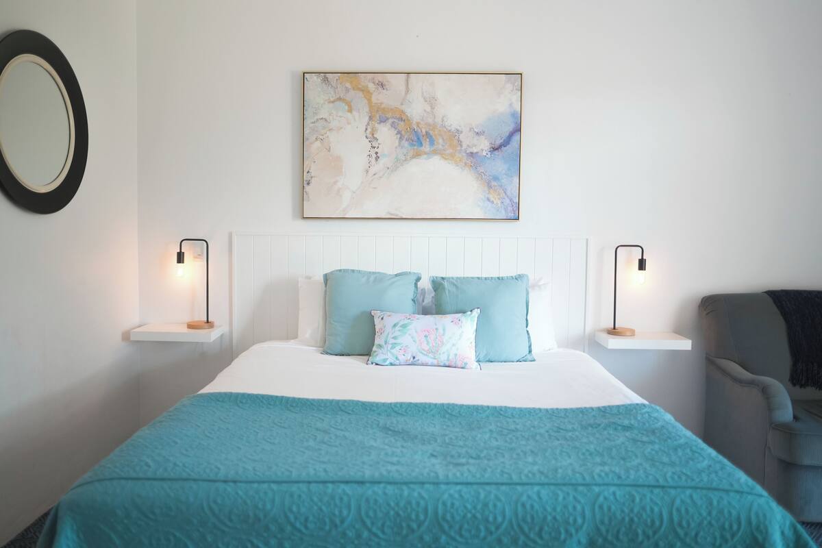 A neatly arranged bedroom with a bed, two bedside tables, a chair, a wall-mounted mirror, and a painting.