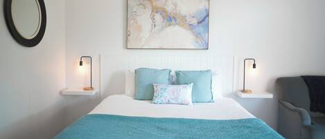 Une chambre à coucher bien rangée, avec un lit, deux tables de chevet, une chaise, un miroir fixé au mur et un tableau.