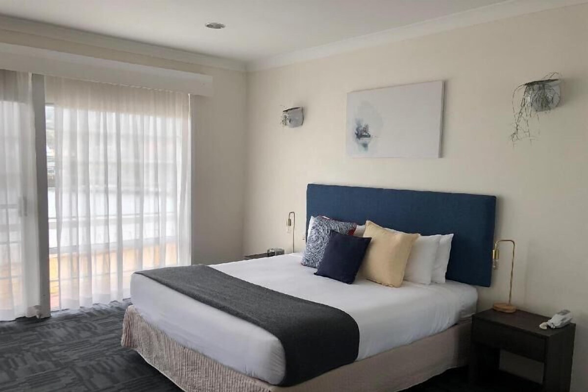 A hotel room with a large bed, a bedside table with a lamp, and a window with sheer curtains.