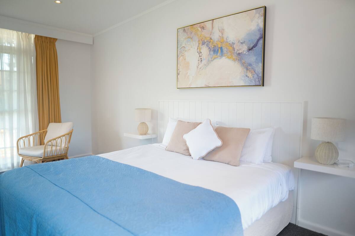 A hotel room with a large bed, a chair, a bedside table, and a wall-mounted abstract painting.