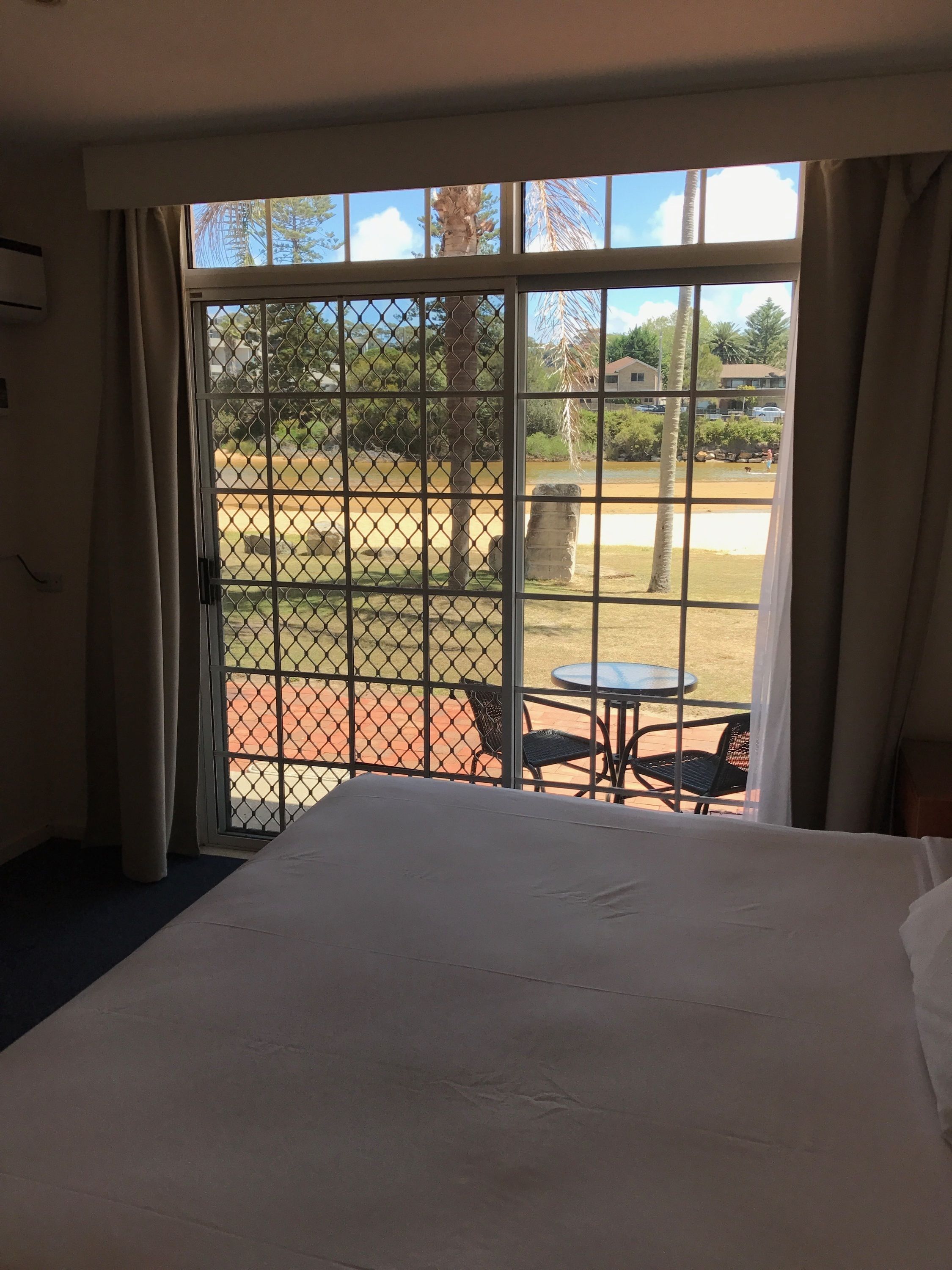 Habitación de hotel con una cama grande, vistas a través de una ventana con celosía metálica y un área exterior con mesa y sillas.