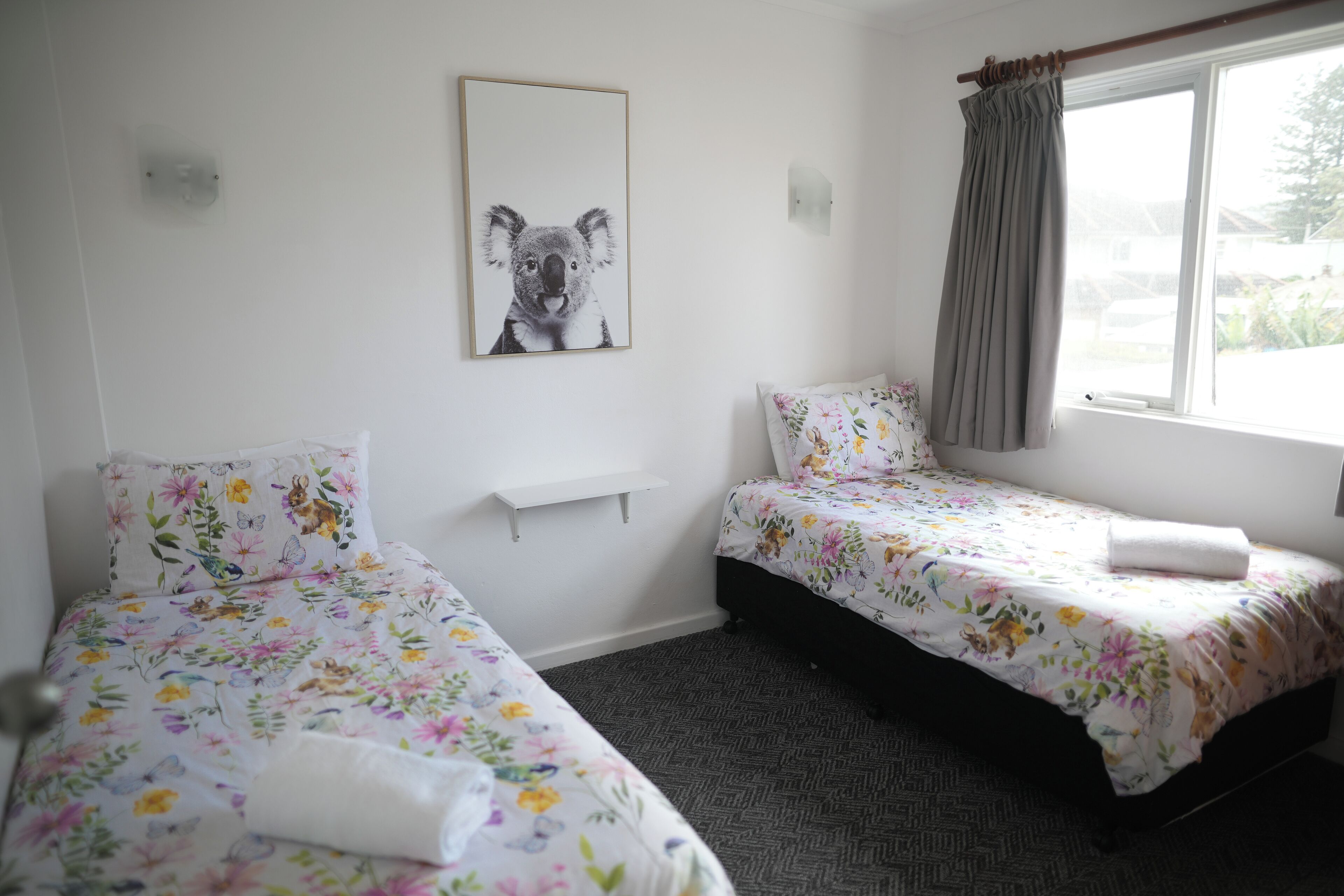 Une chambre avec deux lits simples, une literie à motifs floraux, un tableau représentant un koala accroché au mur, une fenêtre avec des rideaux et une petite étagère.