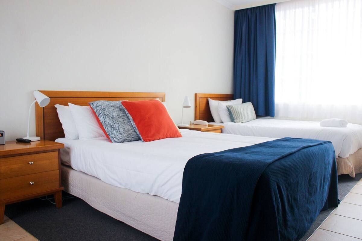 A hotel room with two beds, a wooden headboard, a nightstand with a lamp, and a window with curtains.