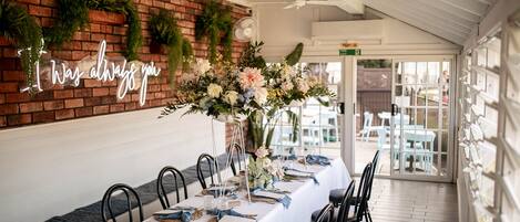 A long dining table with white tablecloths, black chairs, and floral centerpieces. A neon sign on the wall reads "I Was always you."
