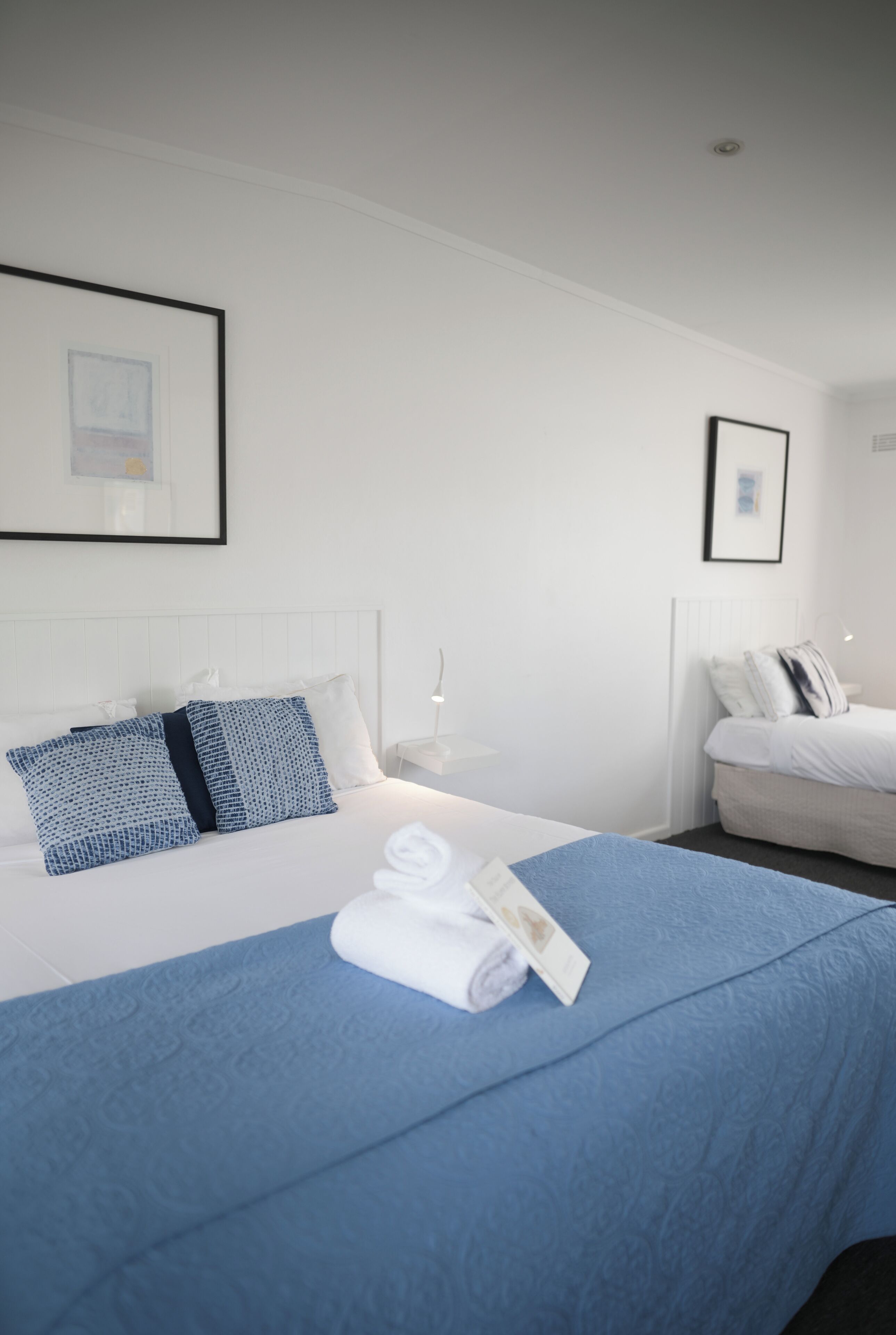 A hotel room with a neatly made bed, a blue blanket, and a rolled towel on the bed.