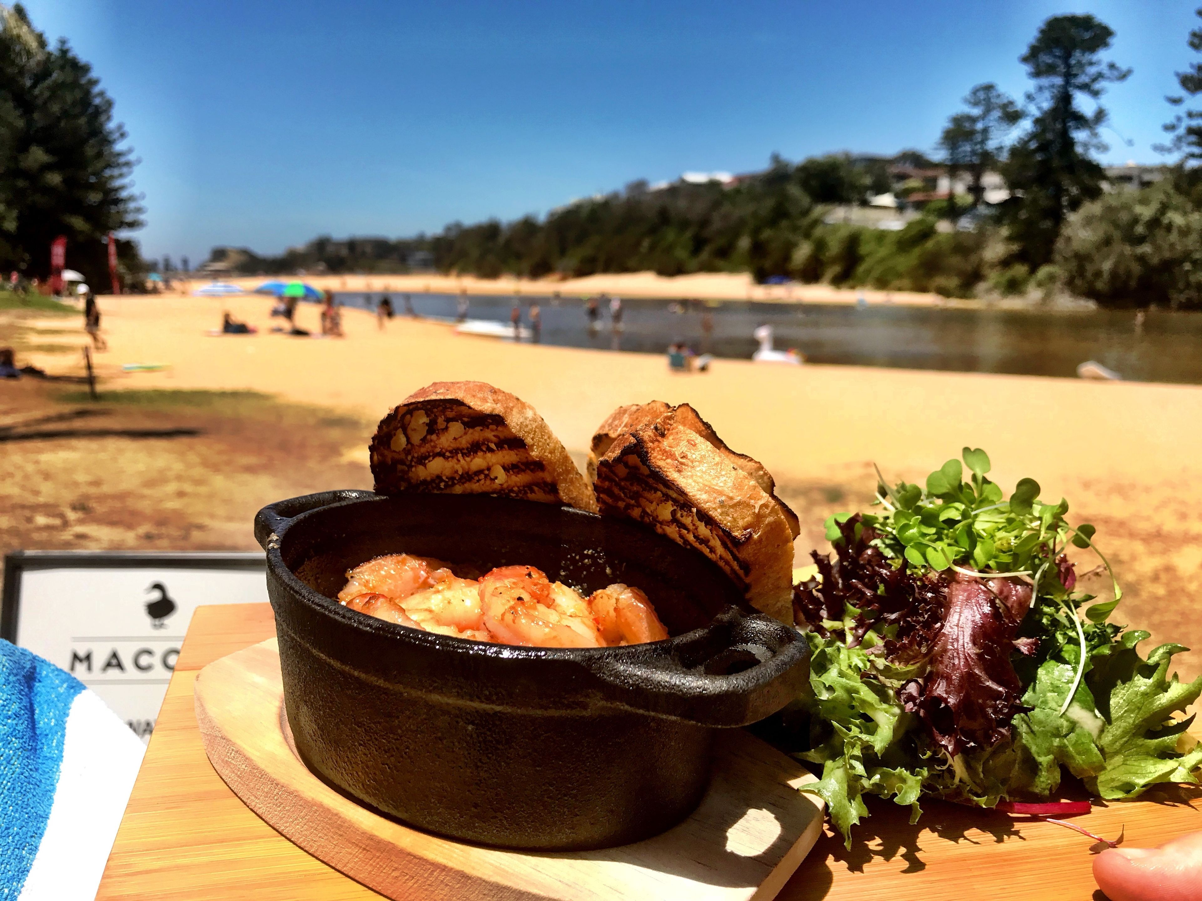 A meal with bread and salad on a wooden board, with a beach and water in the background.