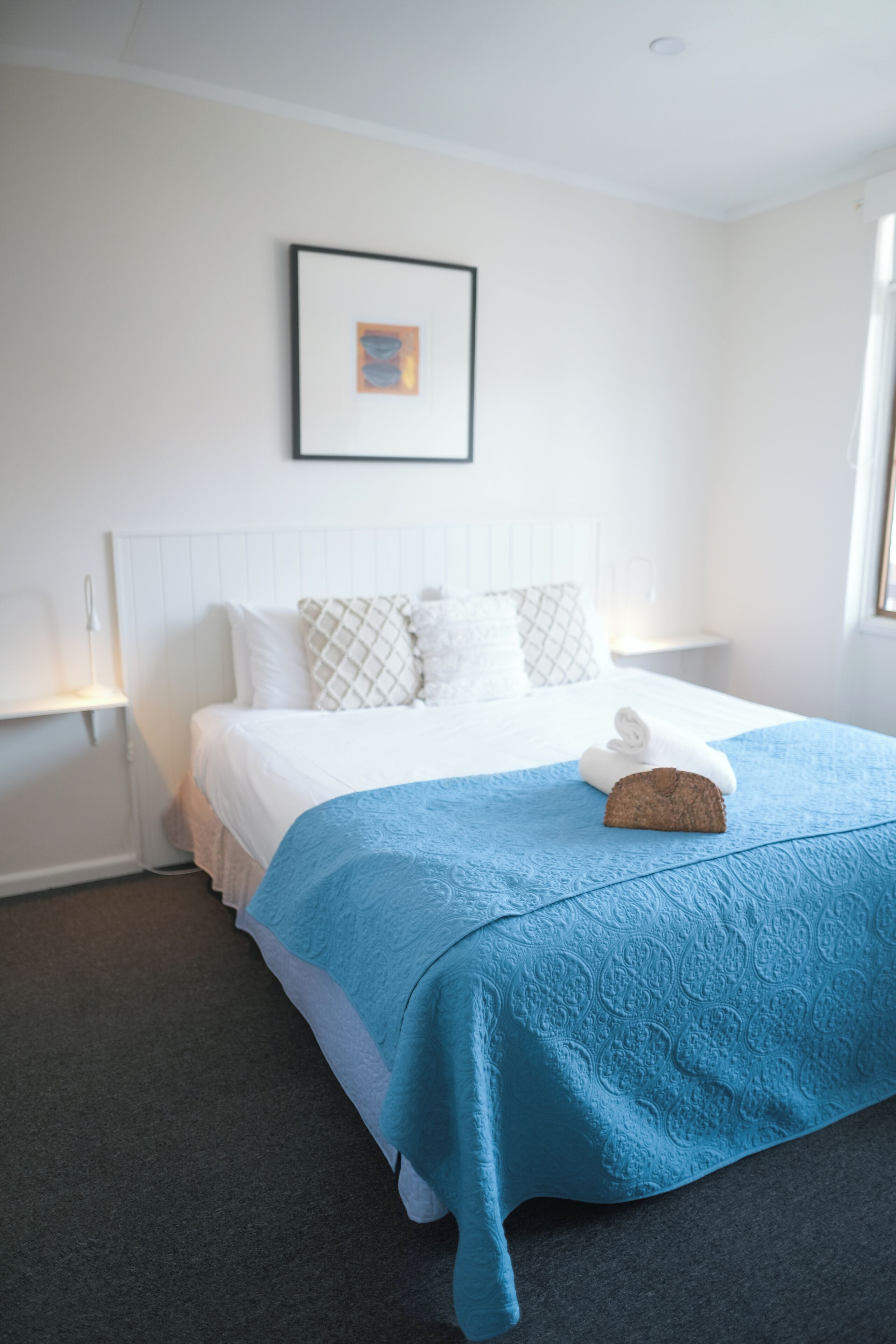 Una cama bien hecha con una colcha azul, almohadas blancas y un cuadro en la pared.