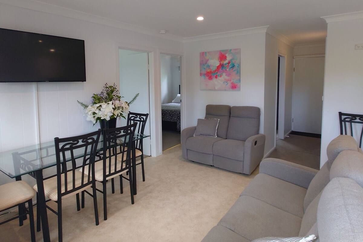 A living room with a grey sofa, a dining table with chairs, a flat-screen TV mounted on the wall, and a painting on the wall.