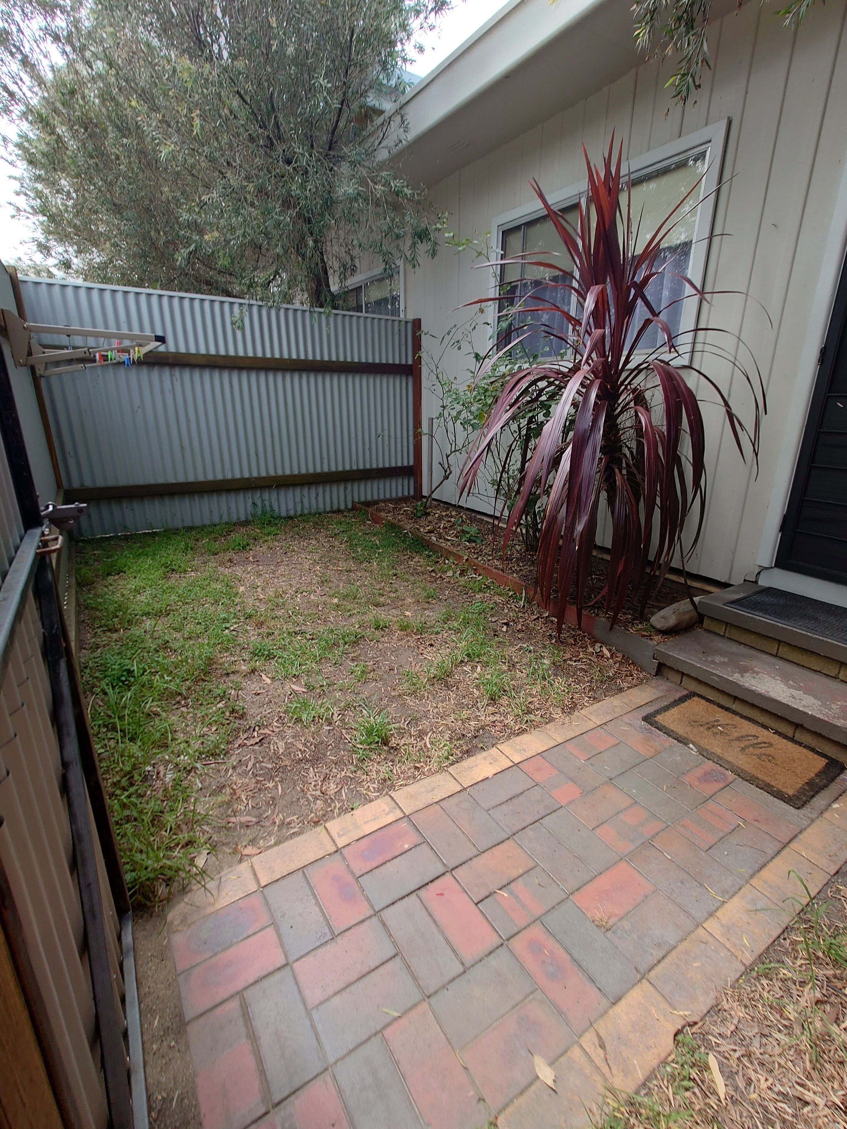 Un jardin avec une terrasse dallée, une clôture en métal et une maison dotée d’une fenêtre.