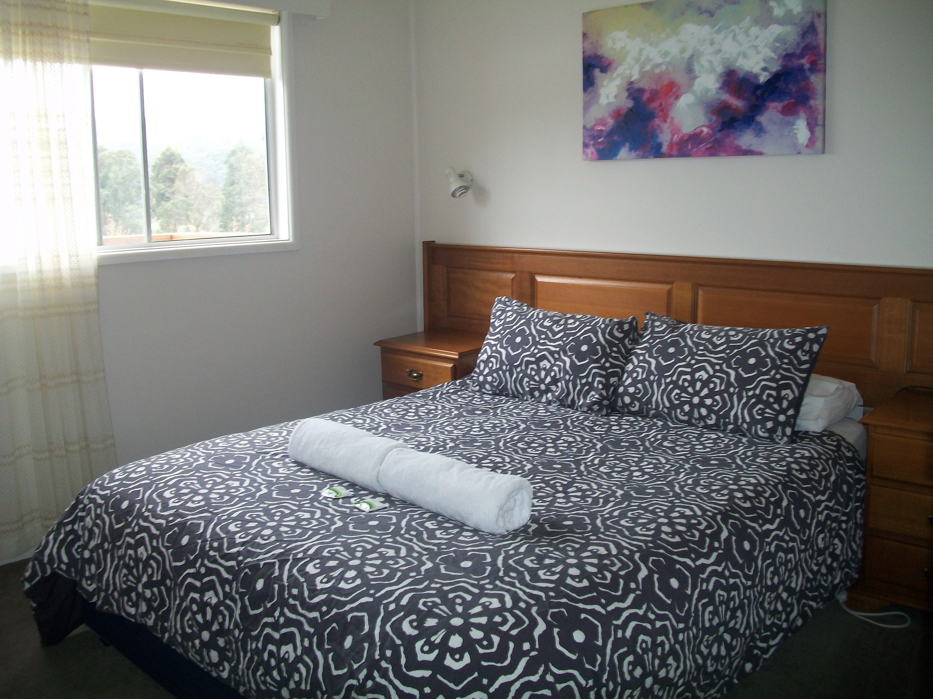 A neatly made bed with a patterned bedspread and pillows, a wooden nightstand with a lamp, and a window with curtains.