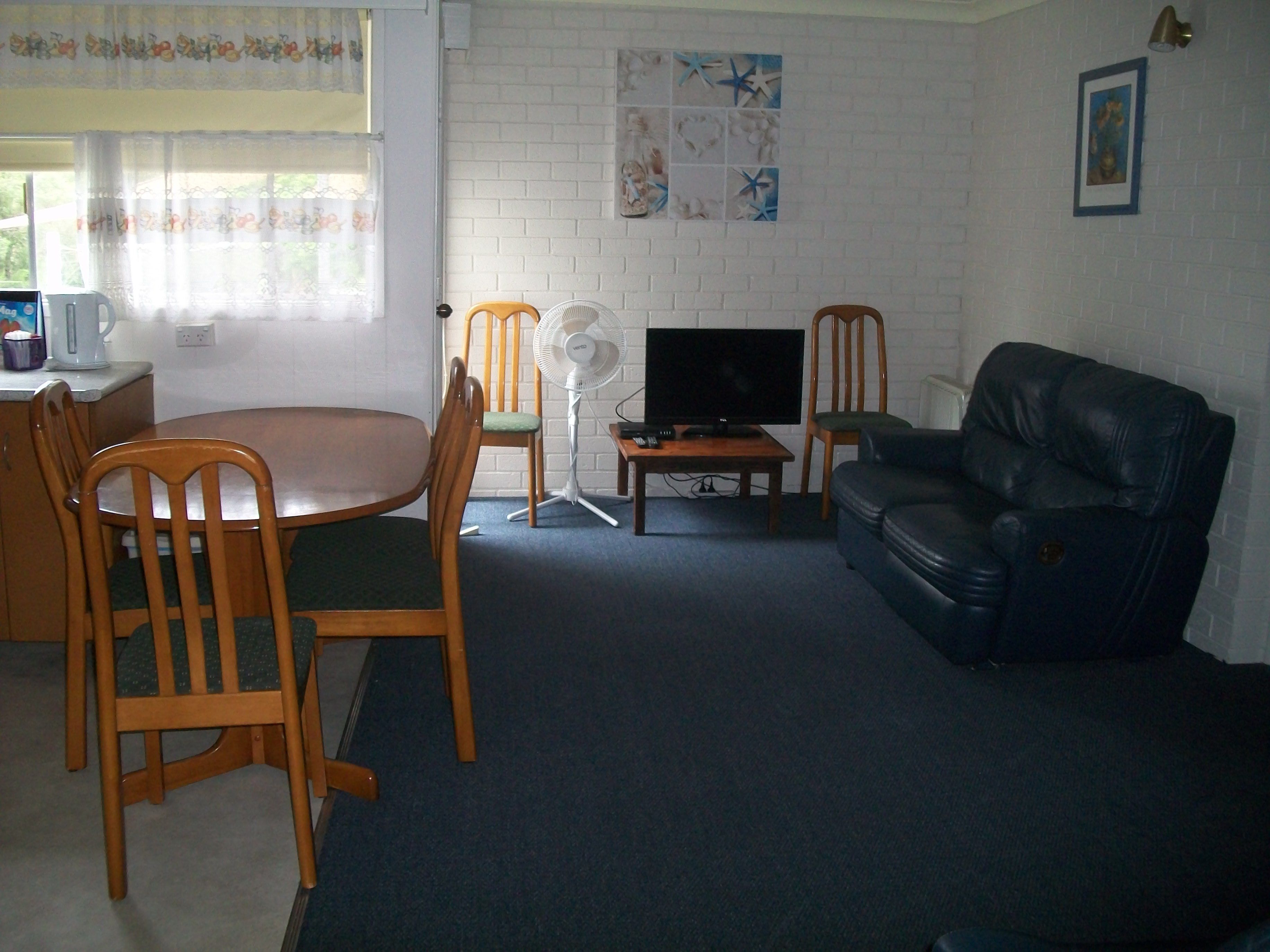 A living room with a dining table, chairs, a black leather sofa, a TV, and a fan.