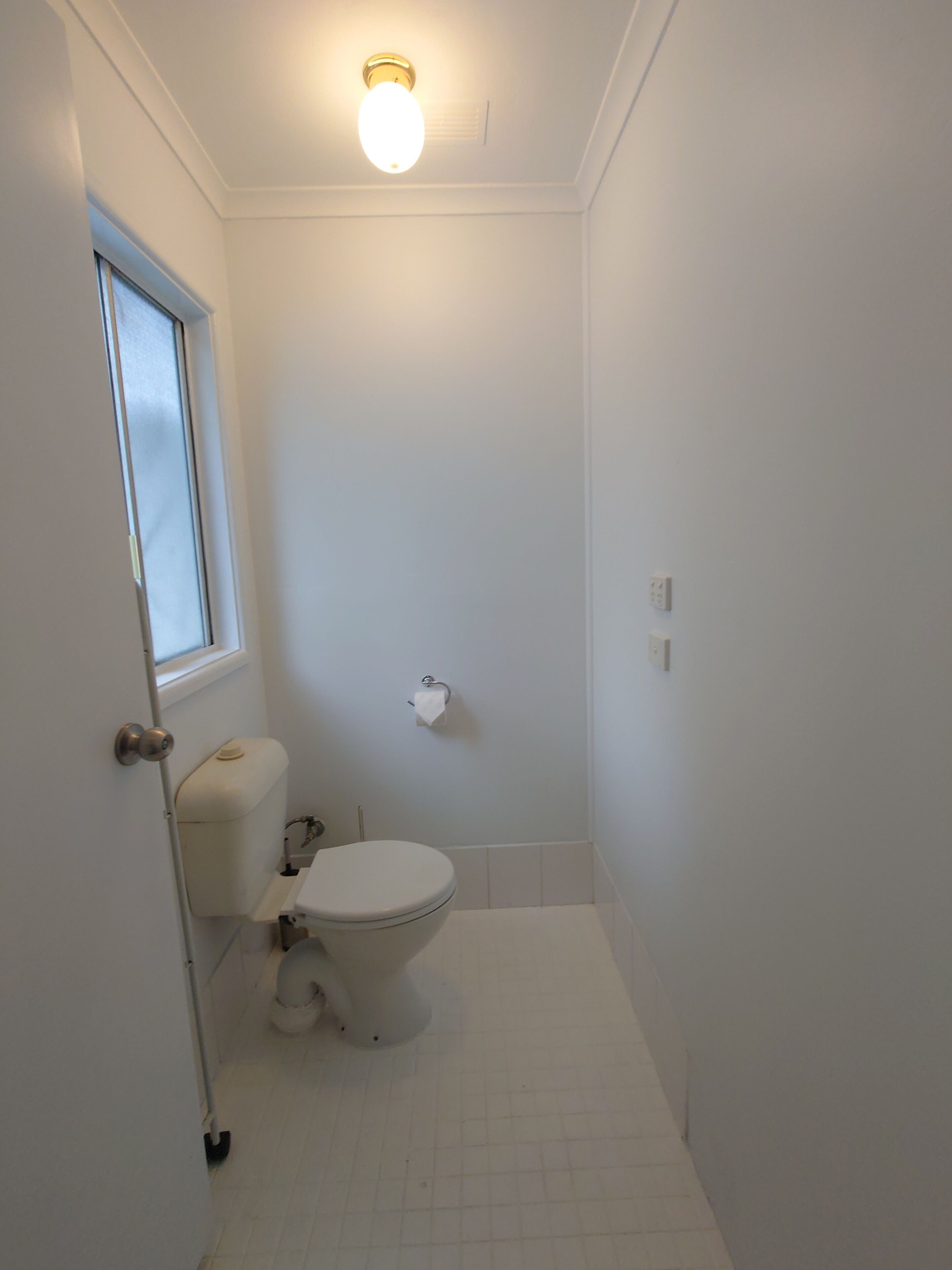 Un baño pequeño y blanco con inodoro, ventana y lámpara en el techo.
