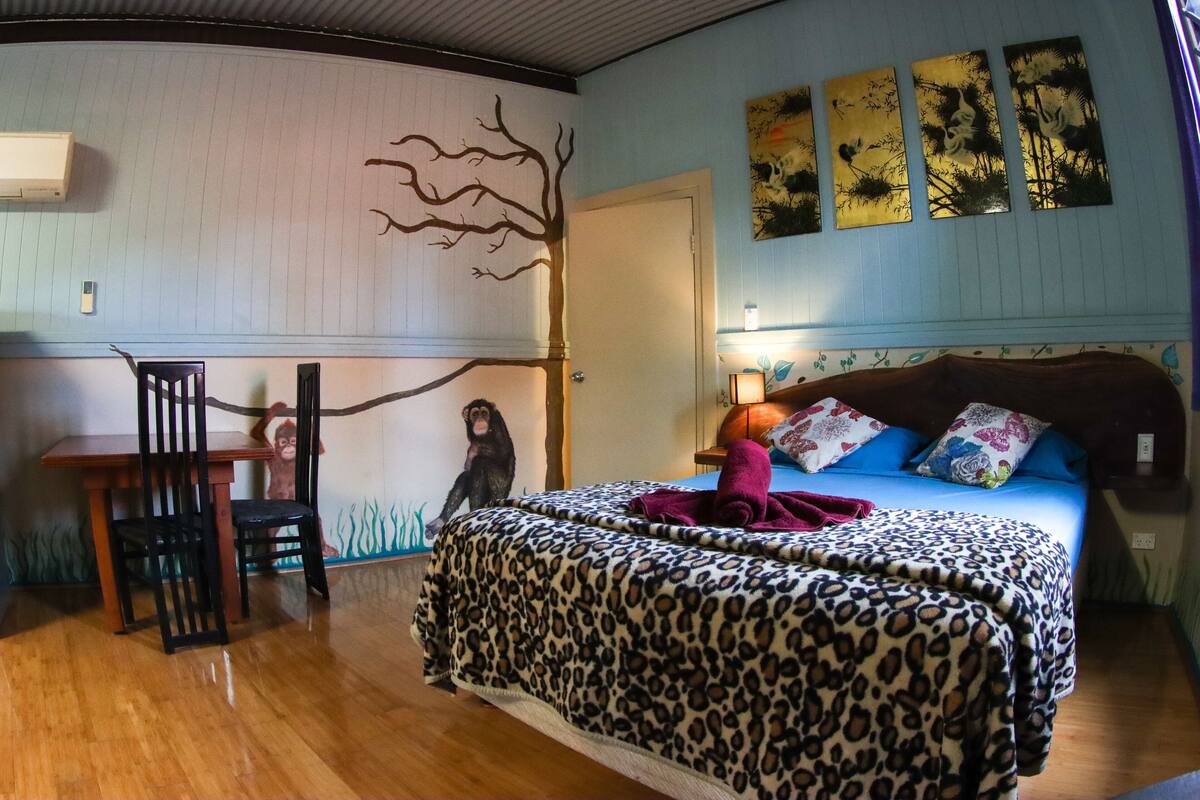 A bedroom with a wooden floor, a bed with a leopard print bedspread, a desk with a chair, and wall art including a tree and three panels.