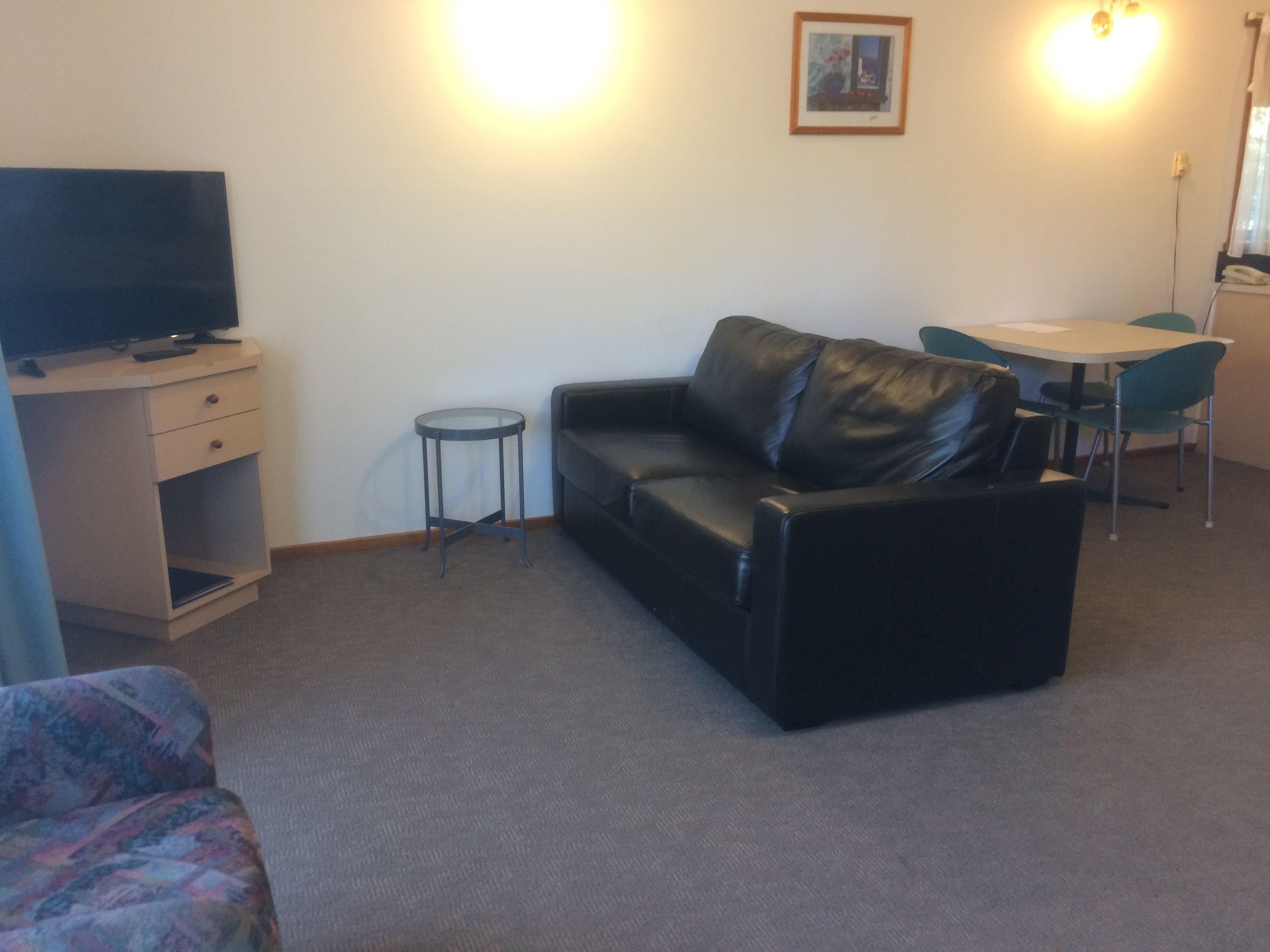 A living room with a black leather sofa, a small round table, a television on a wooden cabinet, and a framed picture on the wall.