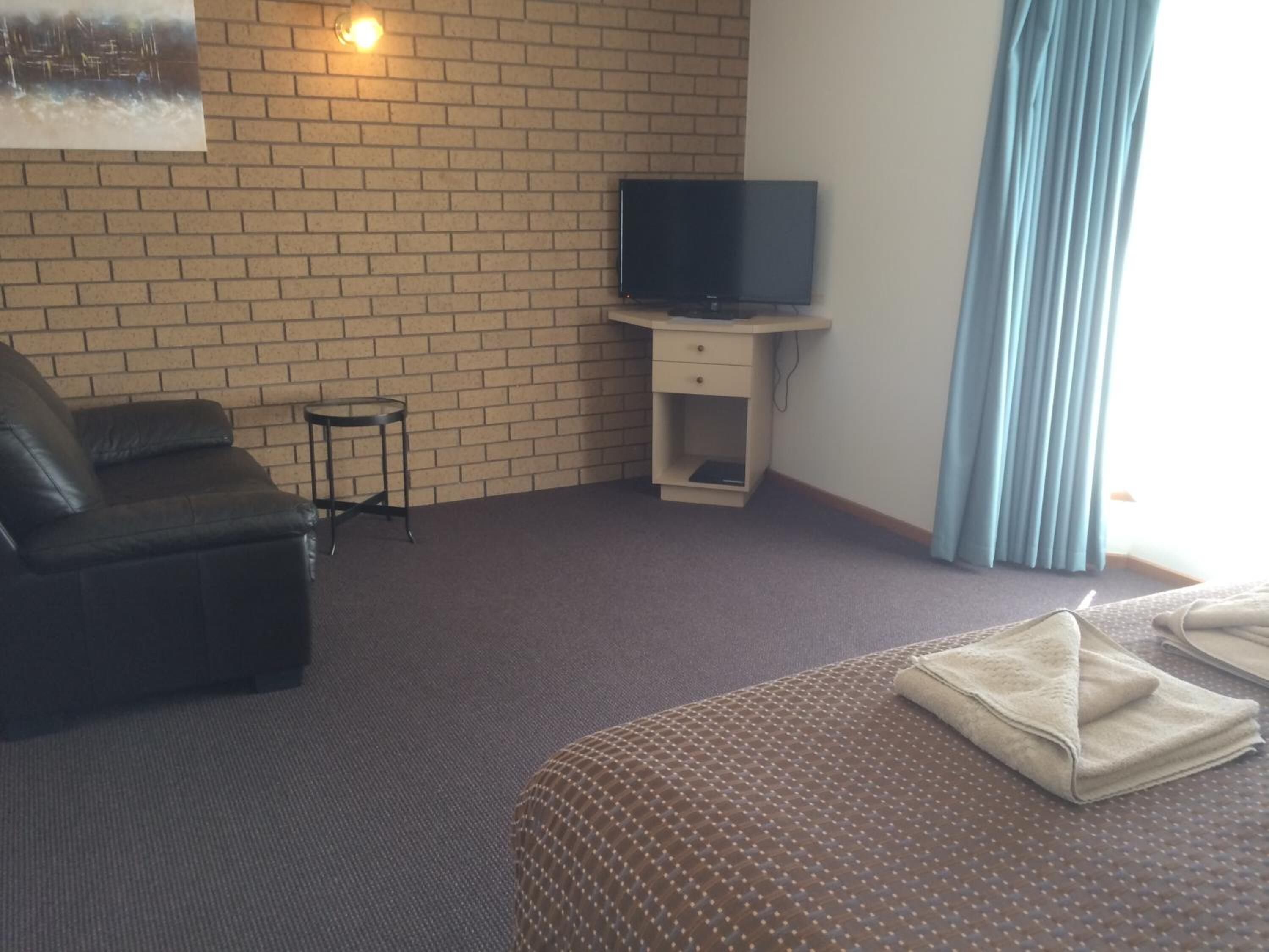 A hotel room with a brick wall, a television on a small table, a leather sofa, a bed with a patterned bedspread, and a window with blue curtains.