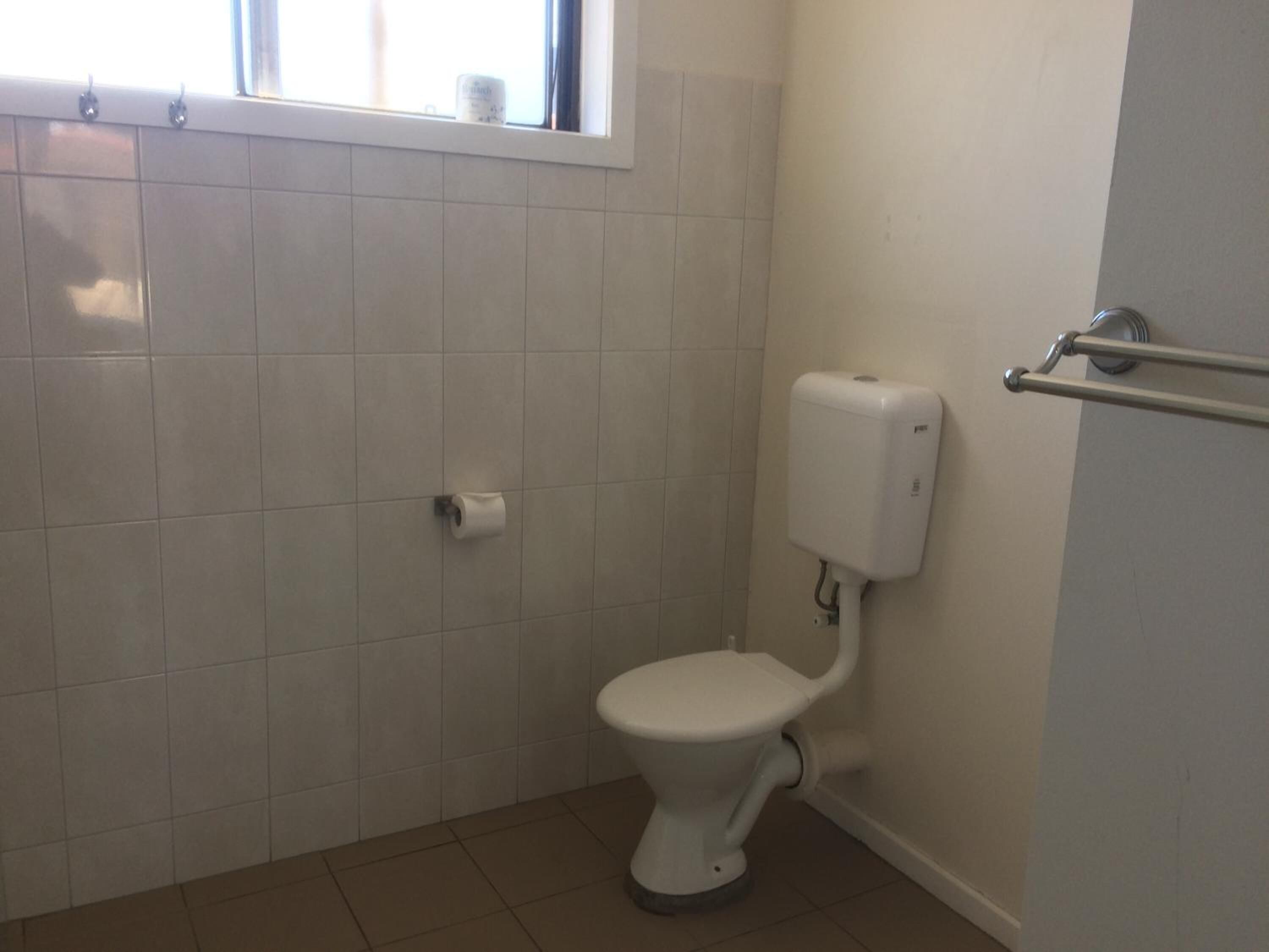 Casa de banho pequena e limpa, com azulejos brancos, sanita, secador de mãos e janela sobre a sanita.