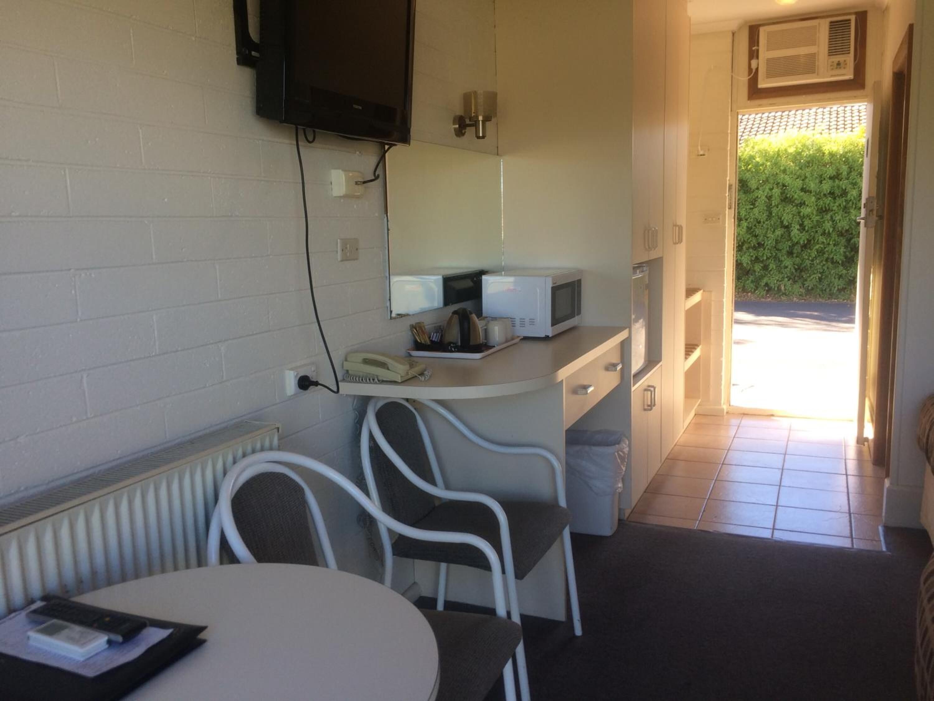 A compact kitchen with a microwave, a telephone, and a small counter. There is a chair and a table with a remote control on it.