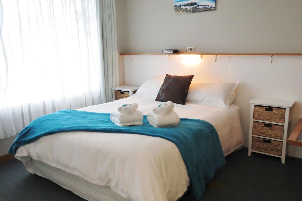 A neatly made bed with a blue blanket, white pillows, and two white towels. There is a small bedside table with a lamp and a wall-mounted picture.
