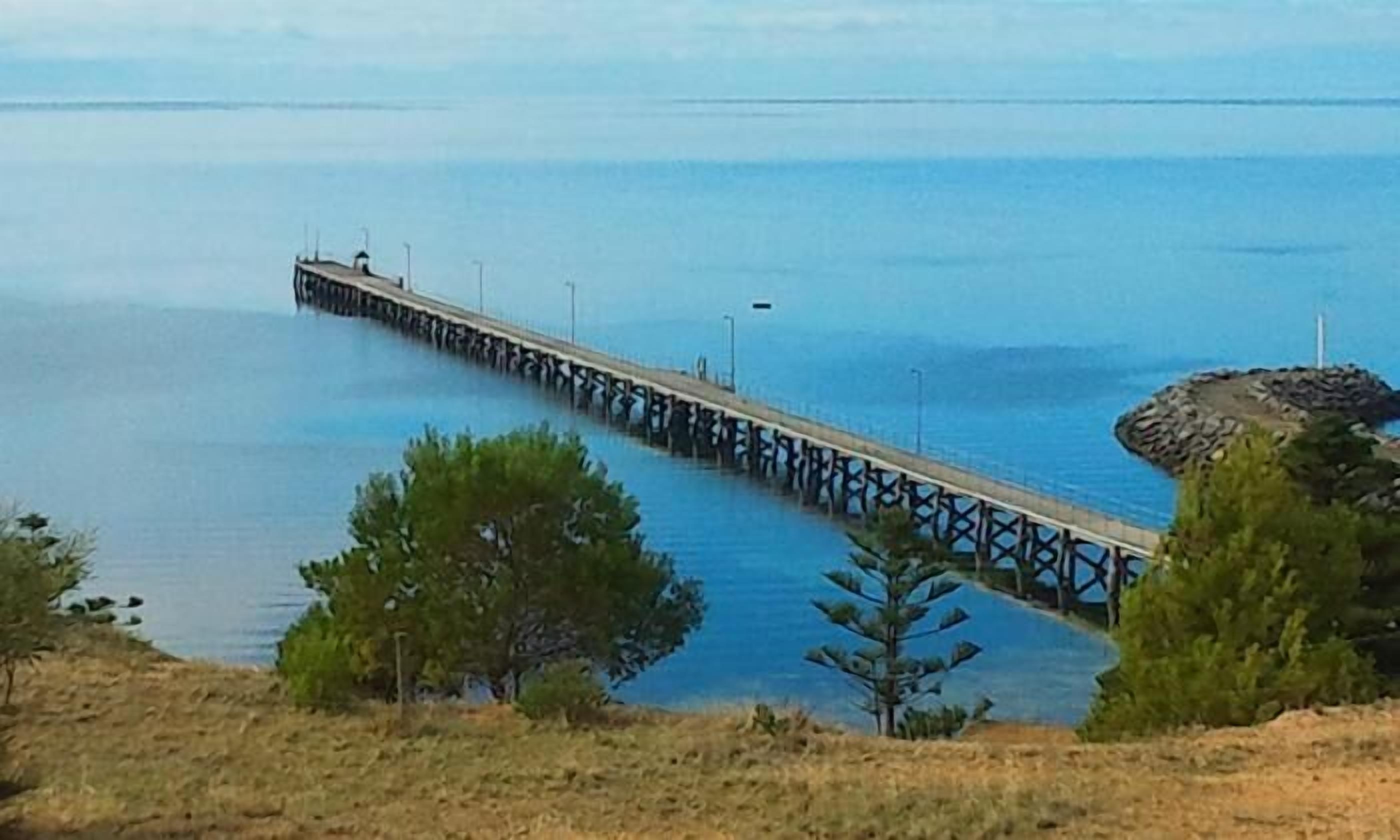 A long pier extending into calm waters, surrounded by sparse vegetation and a rocky outcrop.