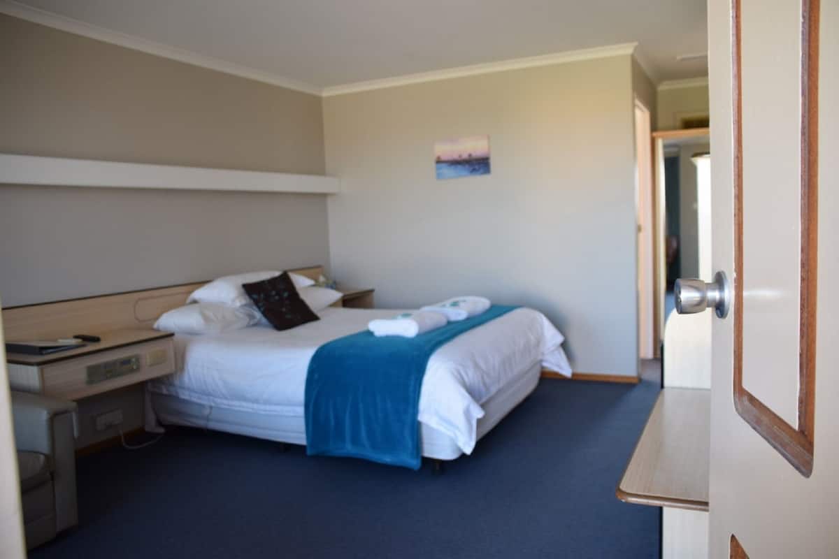 A neatly made bed with a blue blanket, a bedside table with a lamp, and a small painting on the wall.
