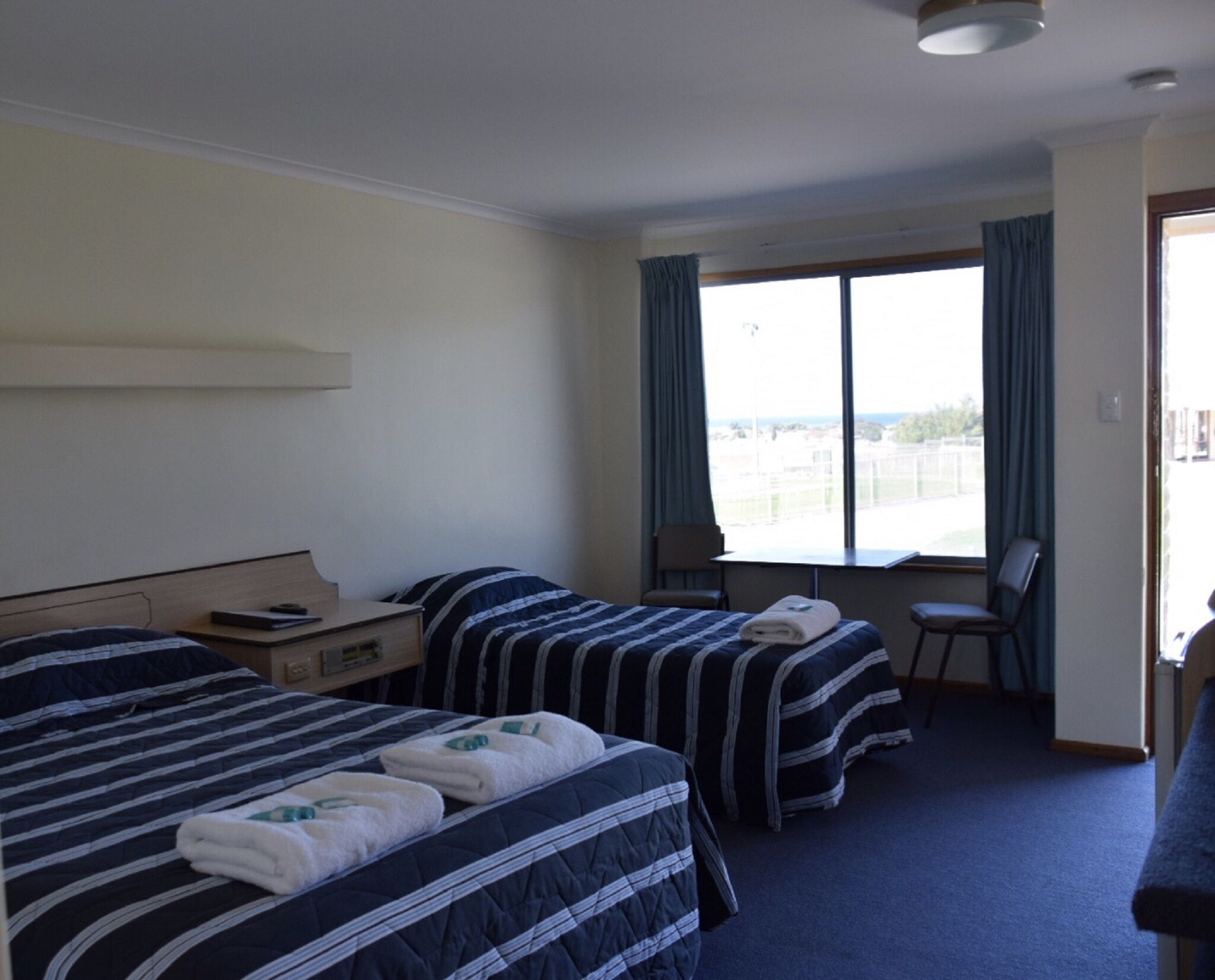 A hotel room with two beds, a desk, a chair, and a large window with curtains.