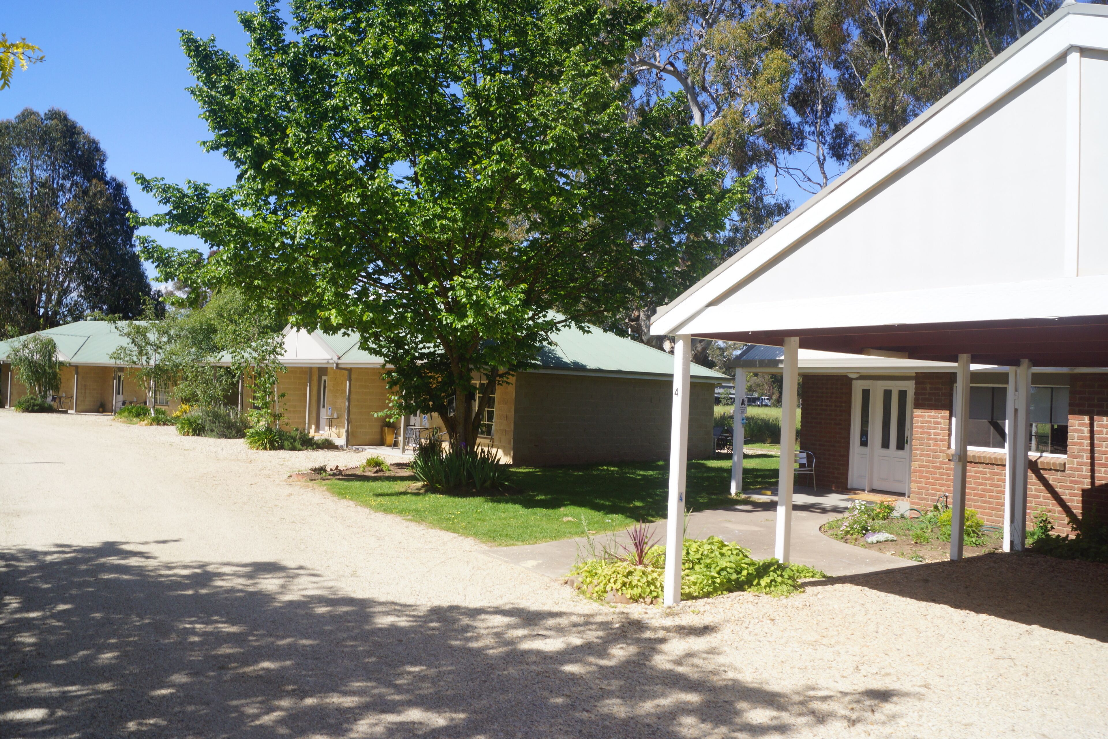 A motel with a covered outdoor seating area, a gravel driveway, and a large tree in front.
