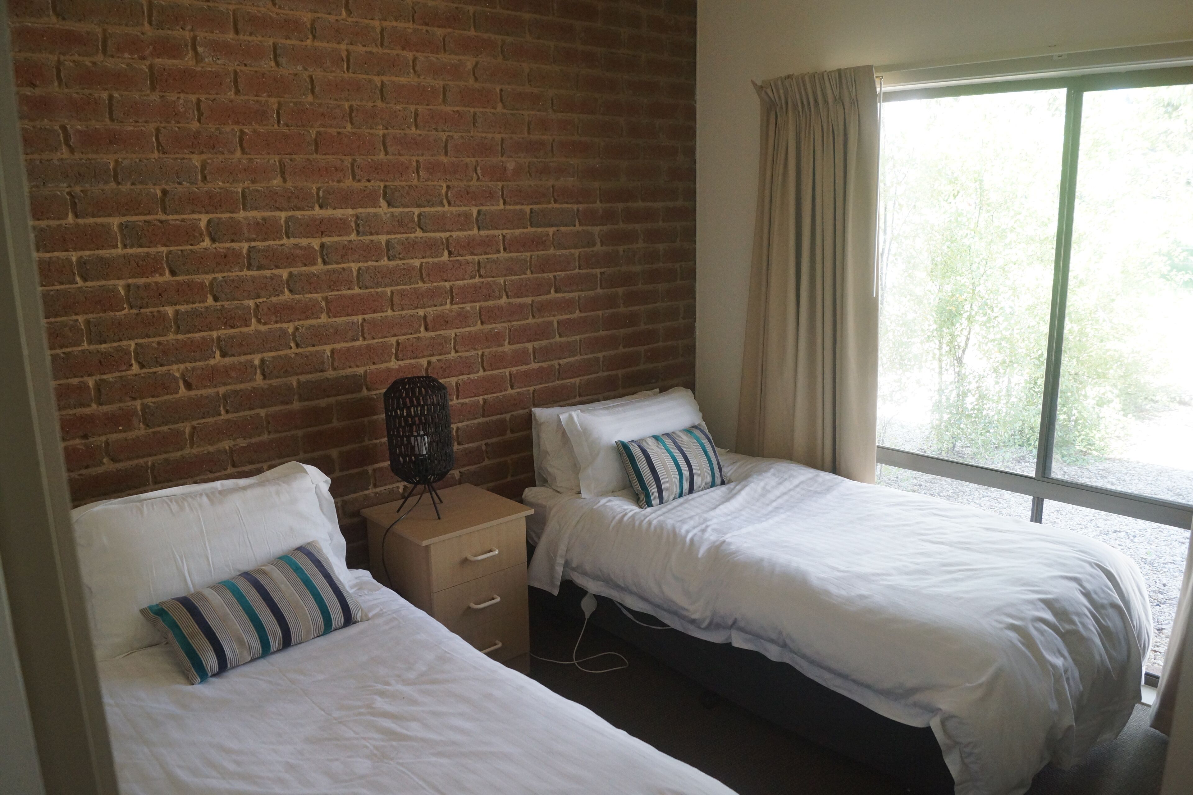 Una habitación con dos camas, una pared de ladrillo, una ventana con cortinas y una pequeña mesa con una lámpara.