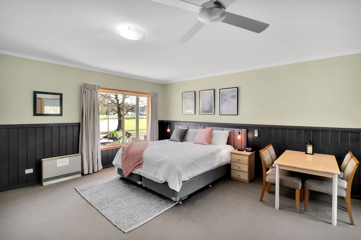 Un dormitorio con cama, mesita de noche, silla y una pequeña mesa con lámpara.