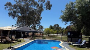 Piscina con sillones de playa y sombrillas, rodeada de árboles y una zona delimitada por una cerca.