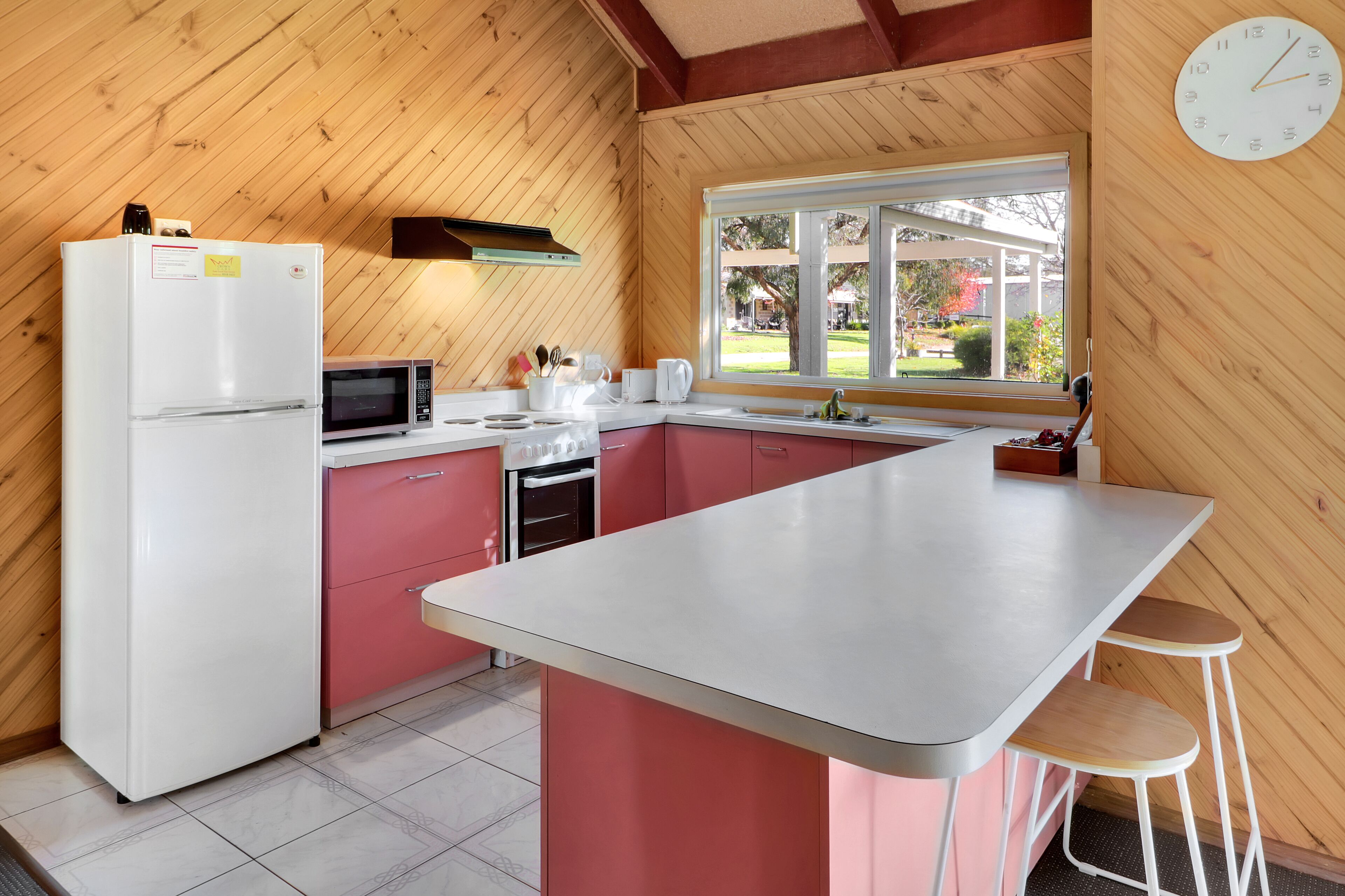 Une cuisine avec des murs en bois, un réfrigérateur blanc, des placards roses, un four à micro-ondes, une machine à café et une fenêtre donnant sur l’extérieur.