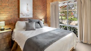 Un dormitorio con pared de ladrillo, un ventanal con cortinas, cama con ropa blanca y una manta gris, y una mesita de noche con lámpara.