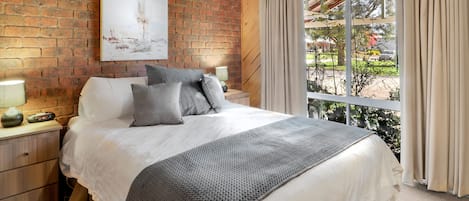 A bedroom with a brick wall, a large window with curtains, a bed with white bedding and a gray blanket, and a nightstand with a lamp.