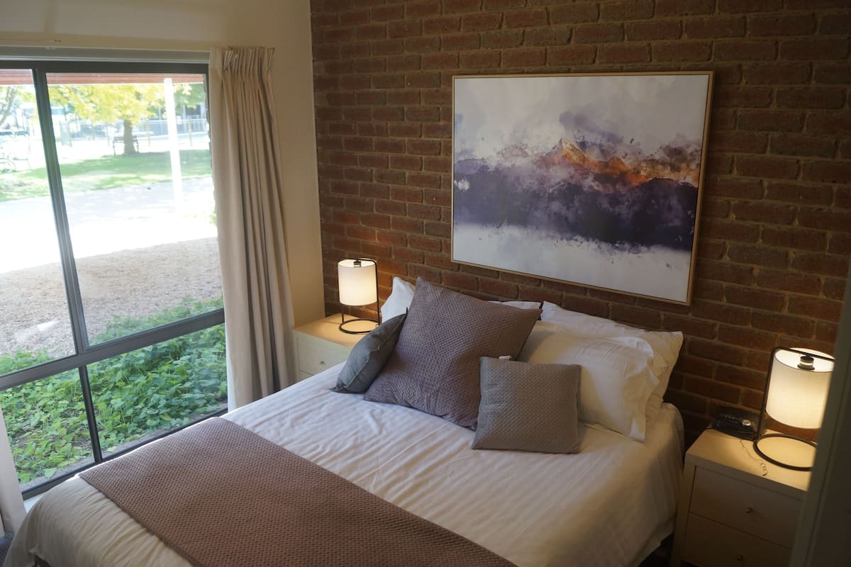 Una cama bien hecha con una manta blanca y marrón, dos lámparas de noche, un cuadro abstracto grande en la pared de ladrillo y vistas a la vegetación a través de un ventanal amplio.