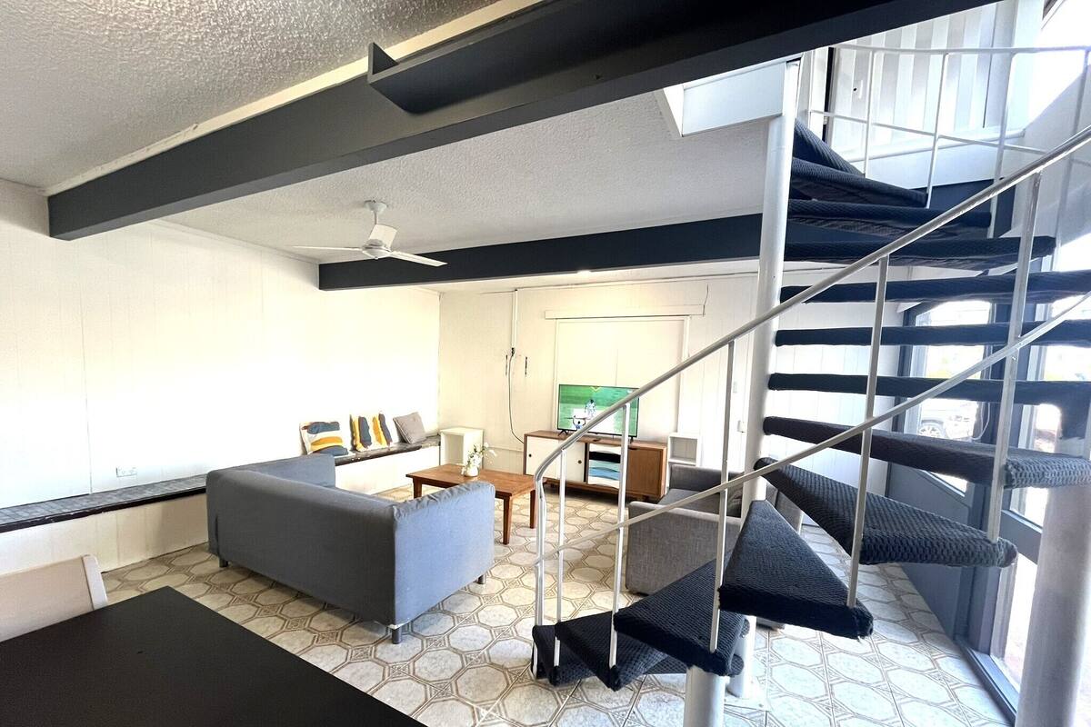 Ein modernes Wohnzimmer mit einer Couch, einem Couchtisch und einem Fernseher. Eine Treppe mit einer Katzenliege und einem Deckenventilator sind ebenfalls vorhanden. Der Raum hat einen Fliesenboden und ein Fenster mit Blick ins Freie.