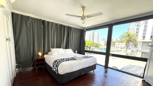 A hotel room with a large bed, a ceiling fan, and a view of the city through large windows. - Tropicana Motel (Mermaid Beach)