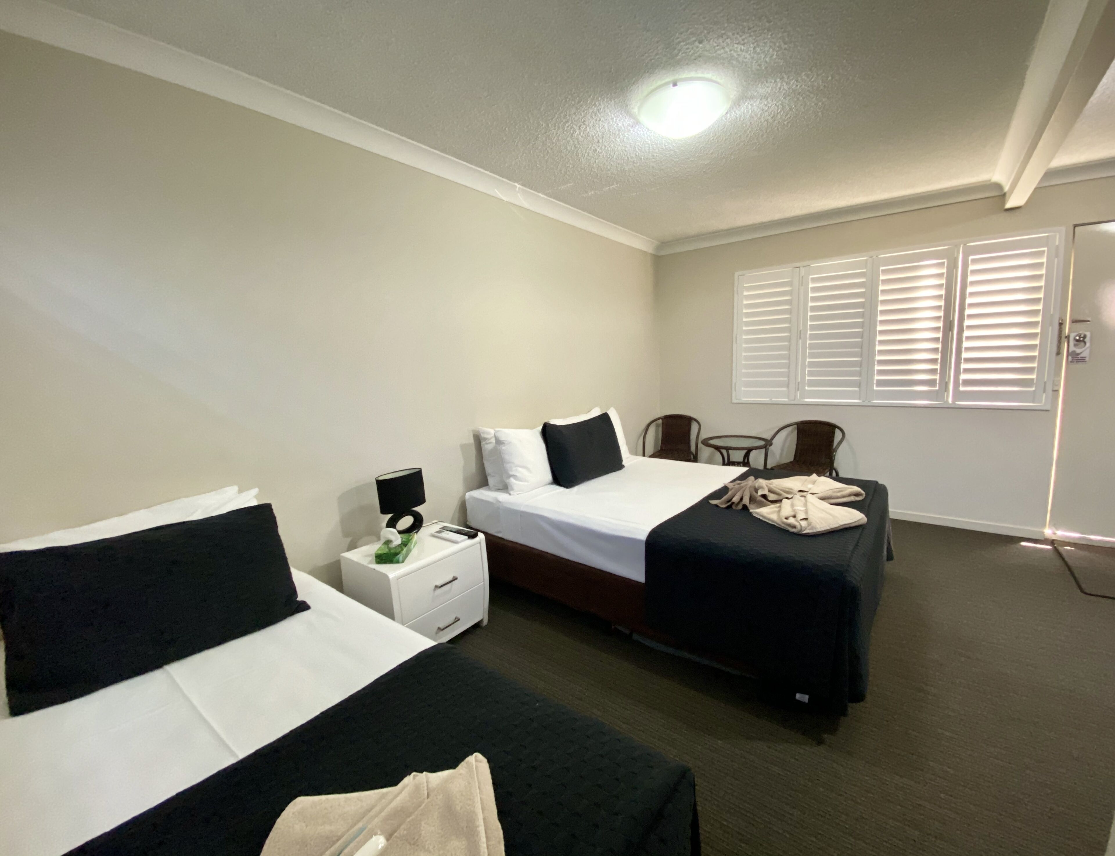Habitación de hotel con dos camas, una mesita con una lámpara, una silla y una ventana con persianas.