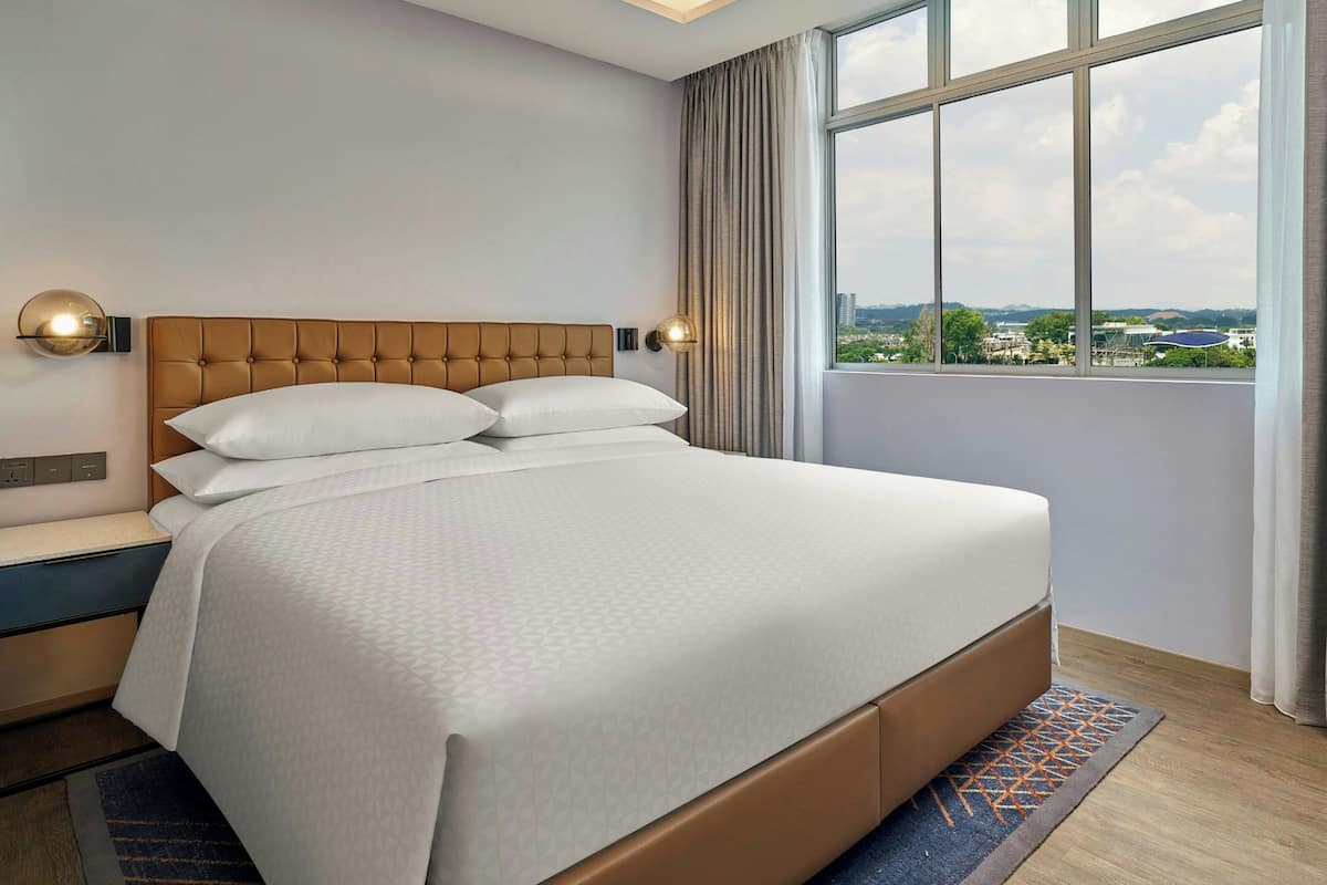 A hotel room with a large bed, a nightstand, a wall-mounted lamp, and a view through a window.