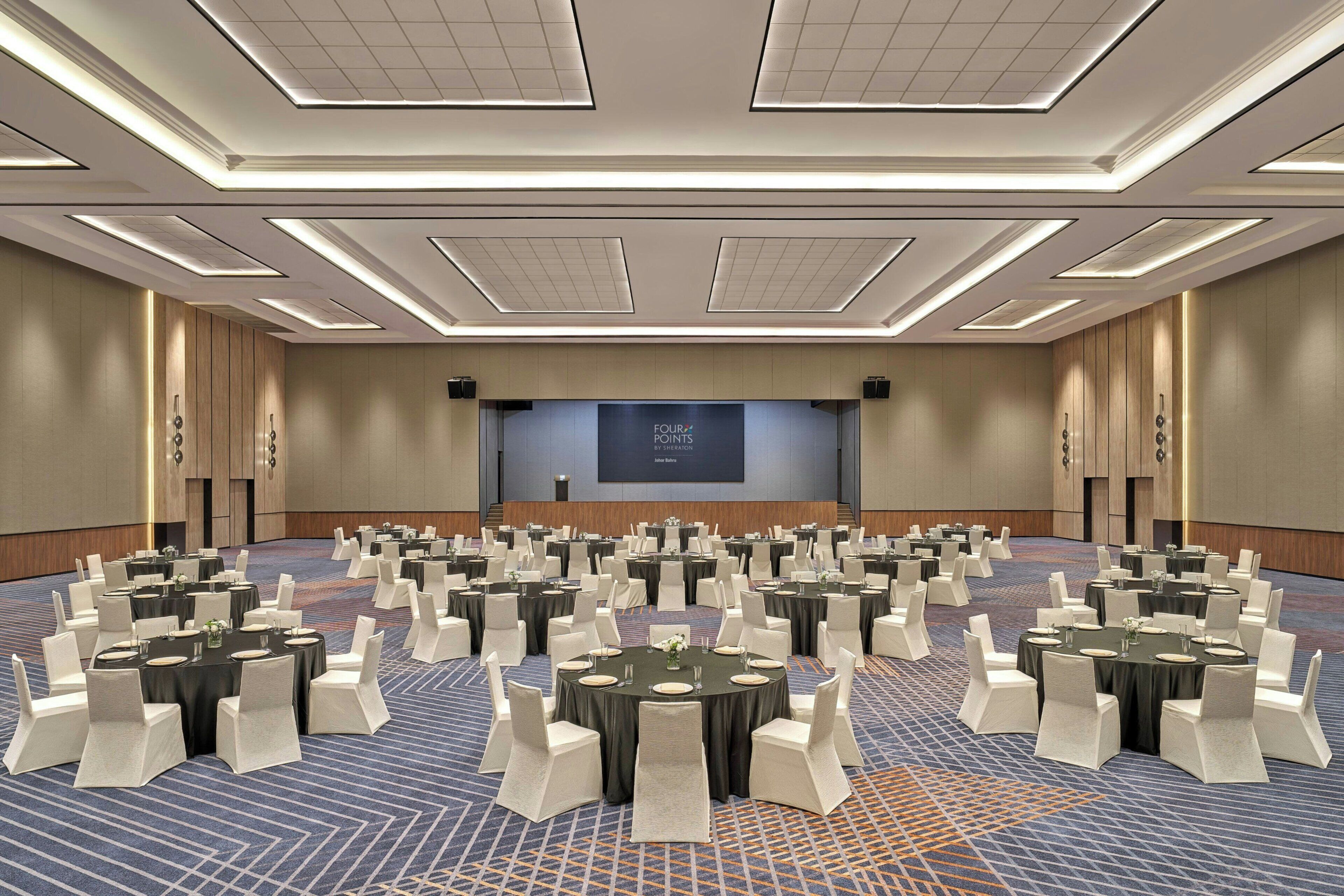 Un salón de conferencias con varias mesas redondas, sillas blancas y una pantalla grande al frente.