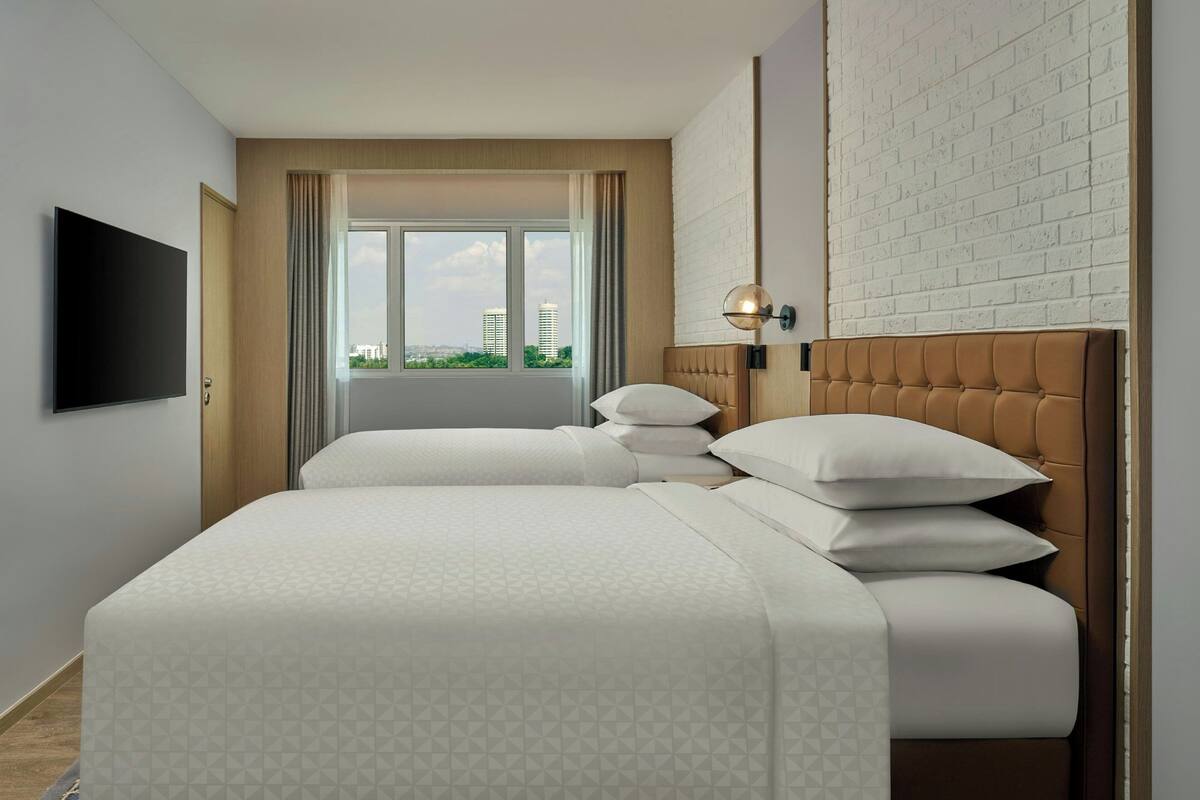A hotel room with a bed, a TV, a window with curtains, and a brick wall.