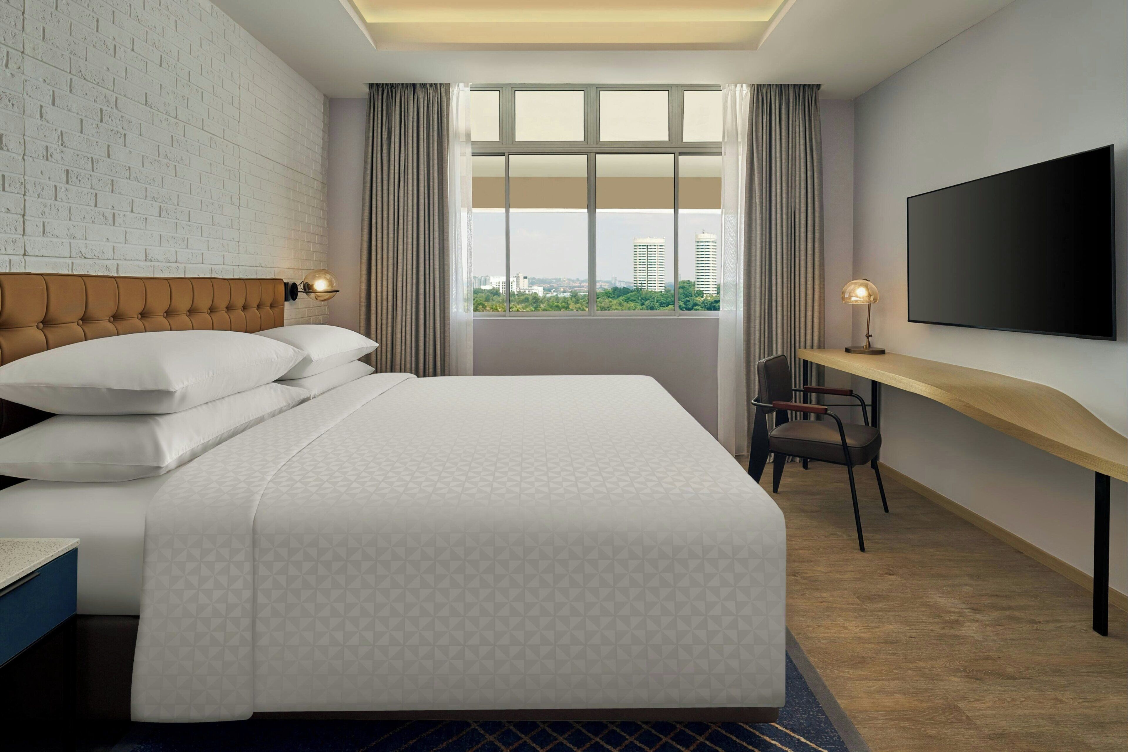 A hotel room with a large bed, a desk, a chair, a TV, and a window with a city view.