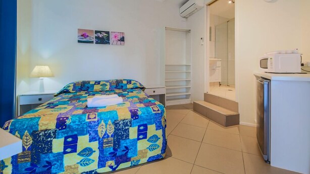 A bedroom with a bed, a nightstand, a lamp, a wall-mounted air conditioner, a small refrigerator, and a bathroom with a shower and toilet. - Marlin Motel (Biggera Waters)