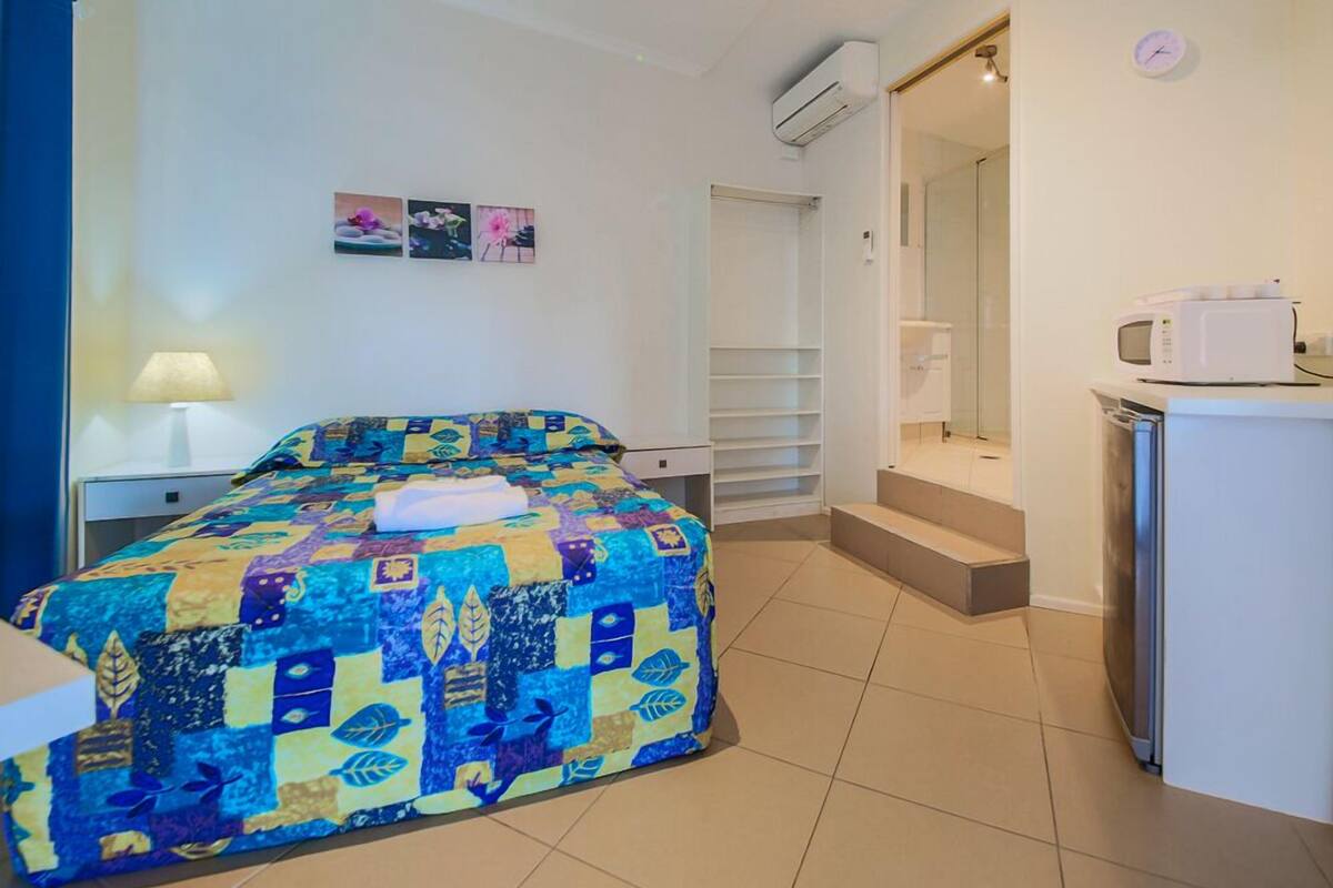 A bedroom with a bed, a nightstand, a lamp, a wall-mounted air conditioner, a small refrigerator, and a bathroom with a shower and toilet.