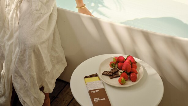 A person in a white bathrobe standing by a bathtub with a plate of strawberries and a chocolate bar on a small round table. - The Hide Byron Bay (Byron Bay)
