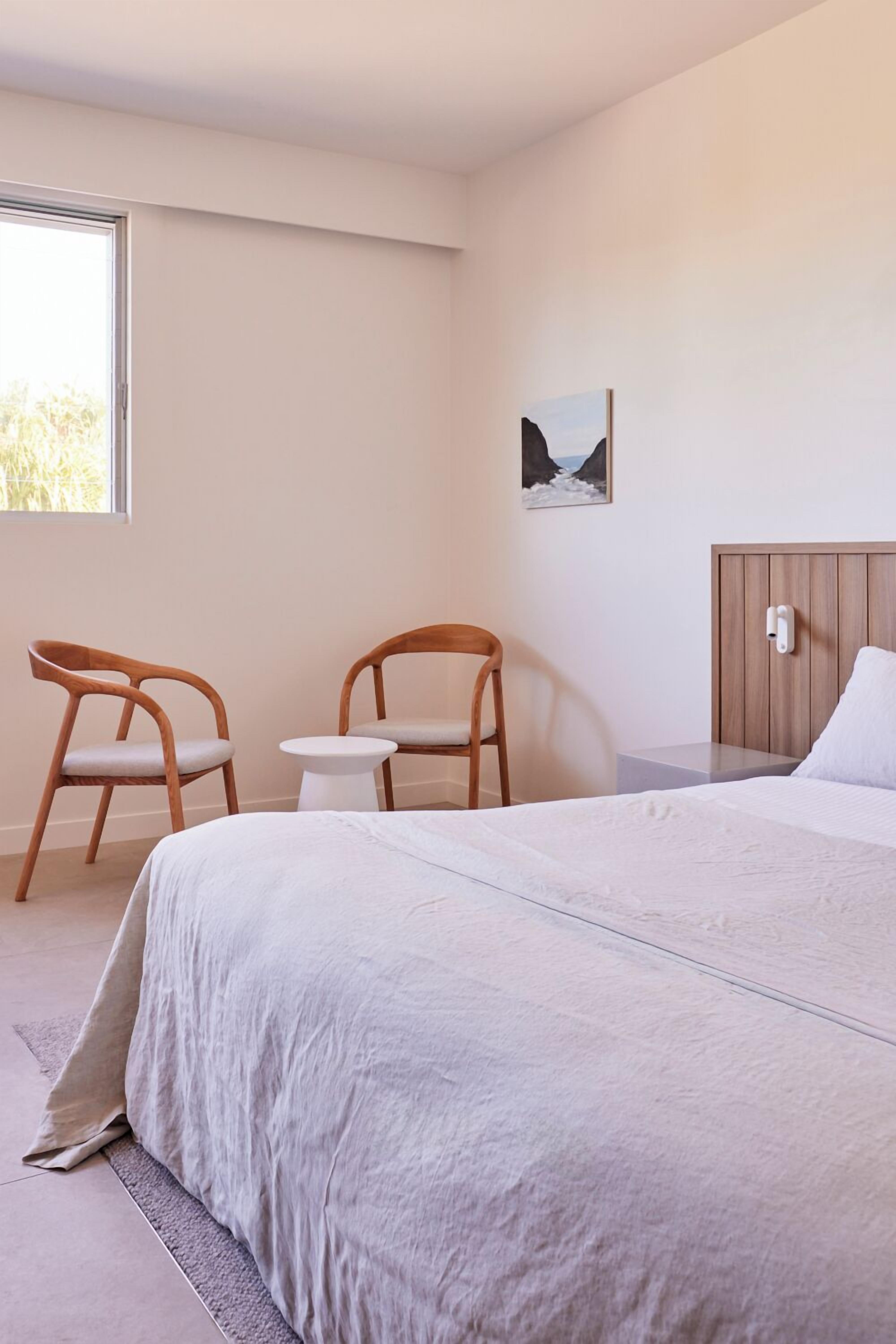 Un dormitorio con una cama, dos sillas de madera, una mesita, una mesita de noche, una lámpara de noche y un cuadro en la pared.