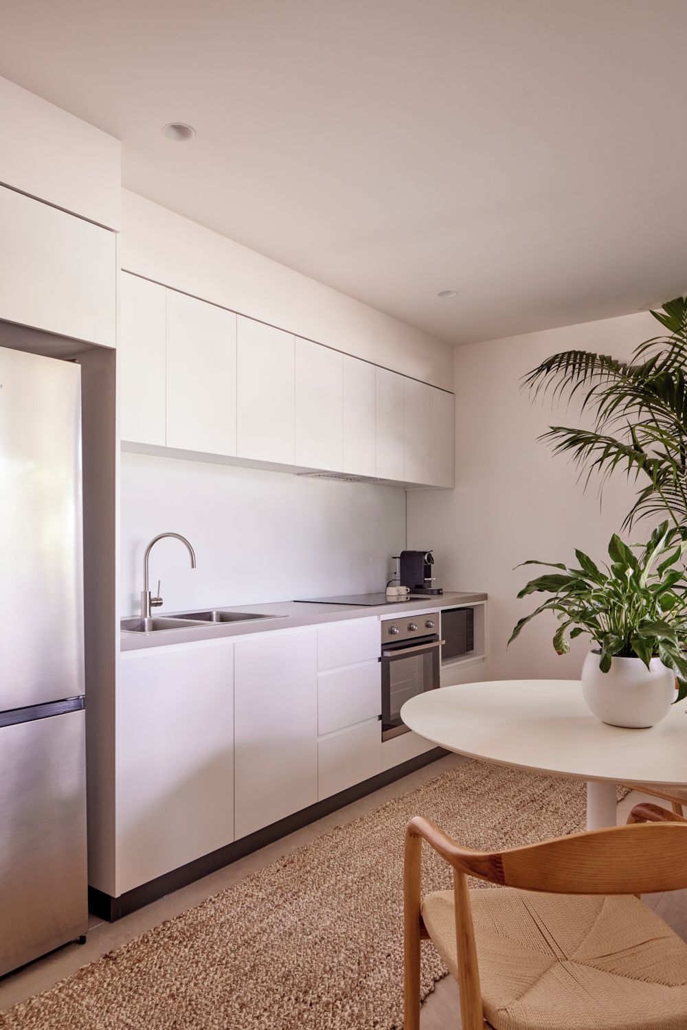 Una cocina moderna con armarios blancos, un refrigerador de acero inoxidable, un lavabo y una cafetera. Hay una mesa redonda con una planta en maceta.