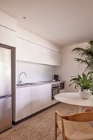 Una cocina moderna con armarios blancos, un refrigerador de acero inoxidable, un lavabo y una cafetera. Hay una mesa redonda con una planta en maceta.